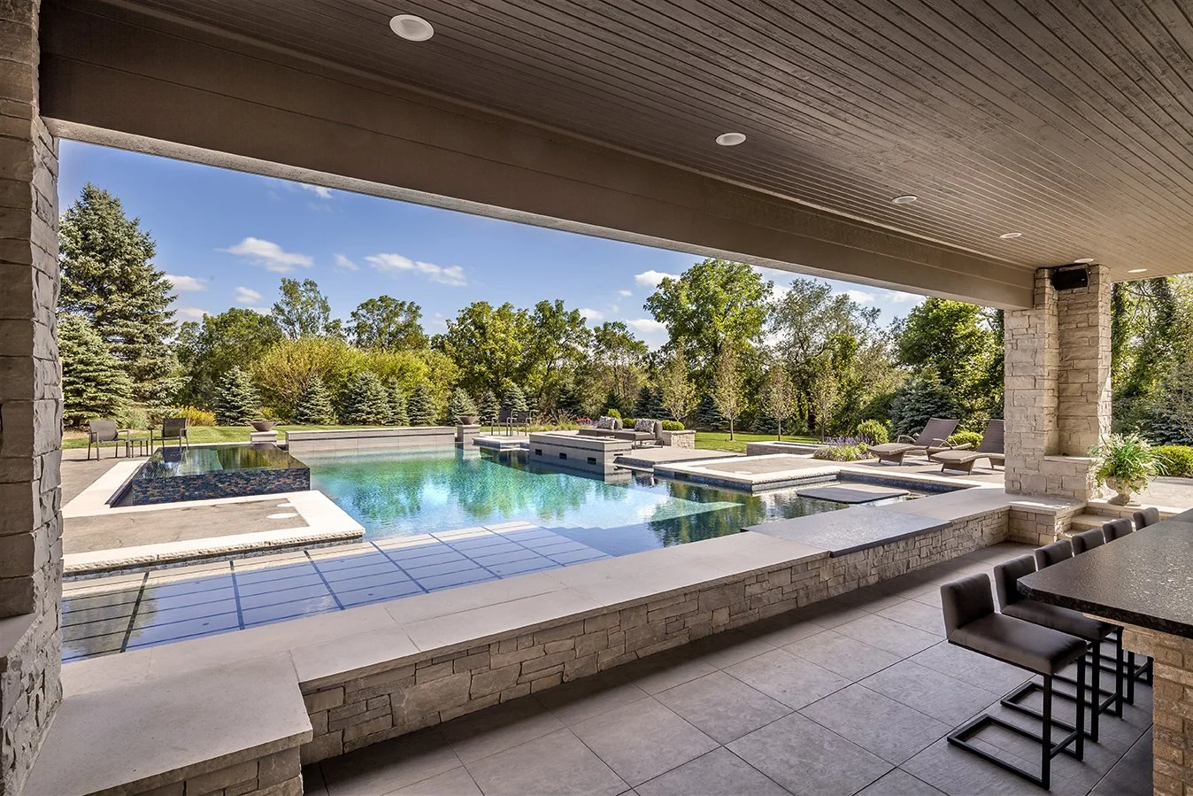 Modern outdoor patio with pool and seating area, surrounded by trees and blue sky.