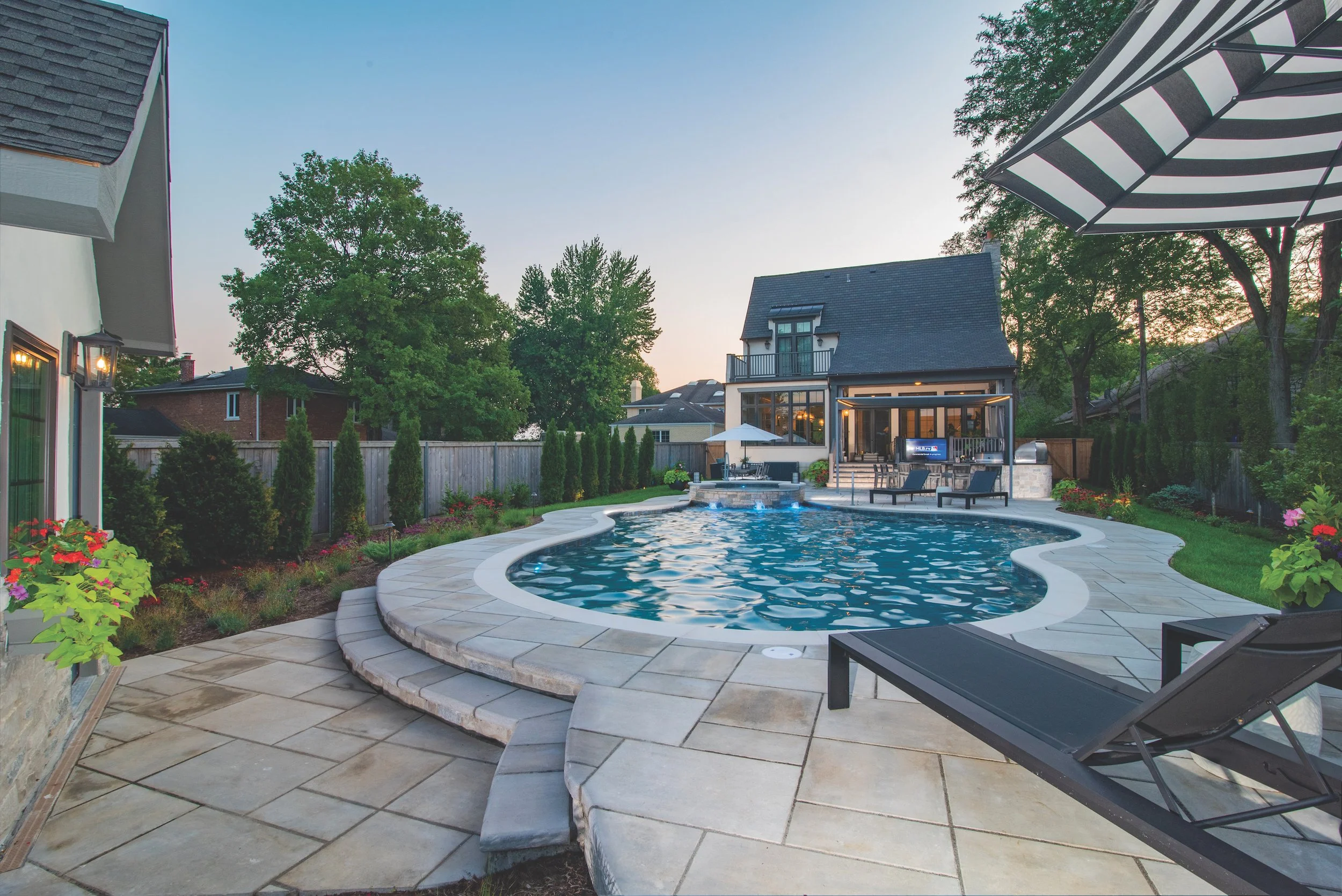 Luxury backyard with swimming pool, patio, lounge chairs, and landscaping, featuring modern house and trees at sunset.