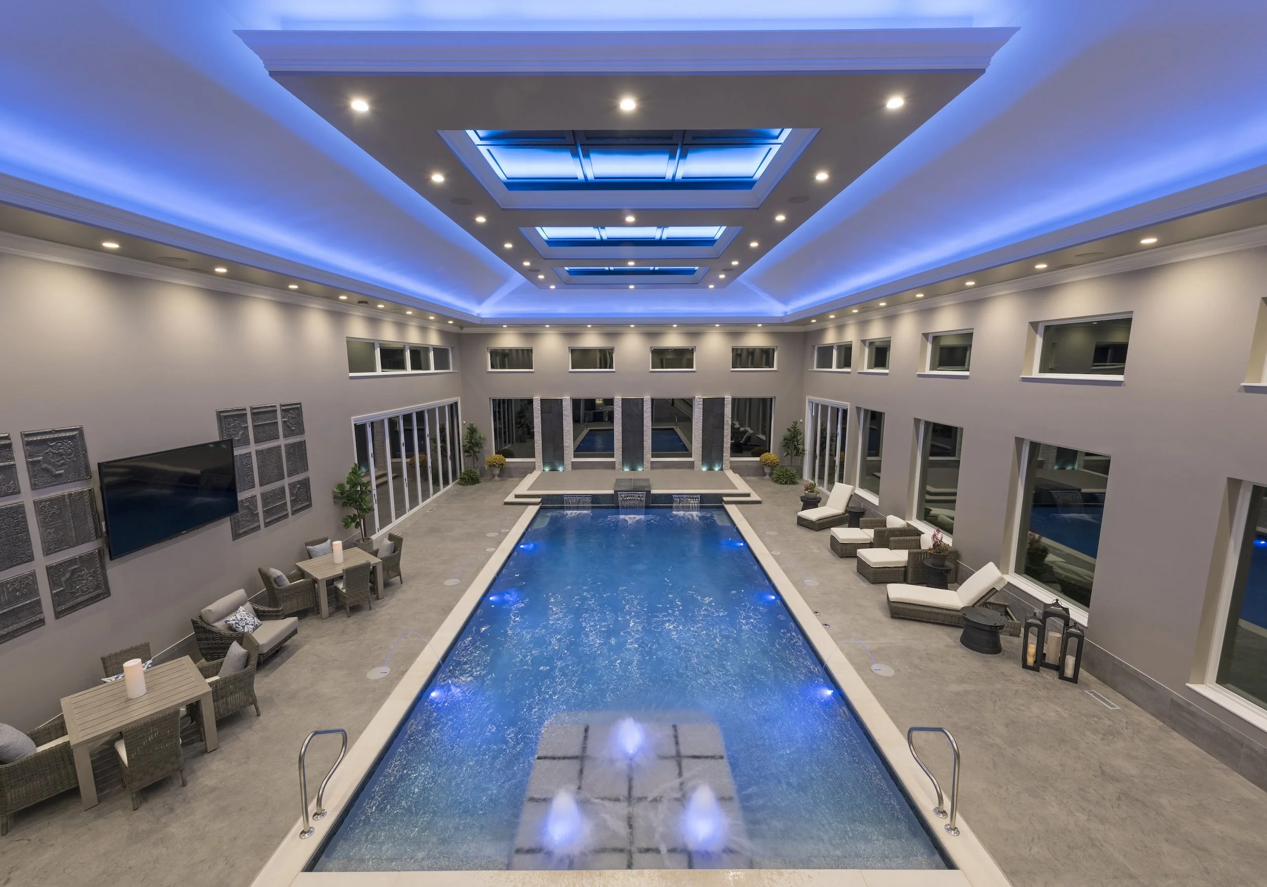 Indoor luxury swimming pool with blue lighting, surrounded by modern lounge chairs and tables, featuring skylights on the ceiling and large windows on the walls.