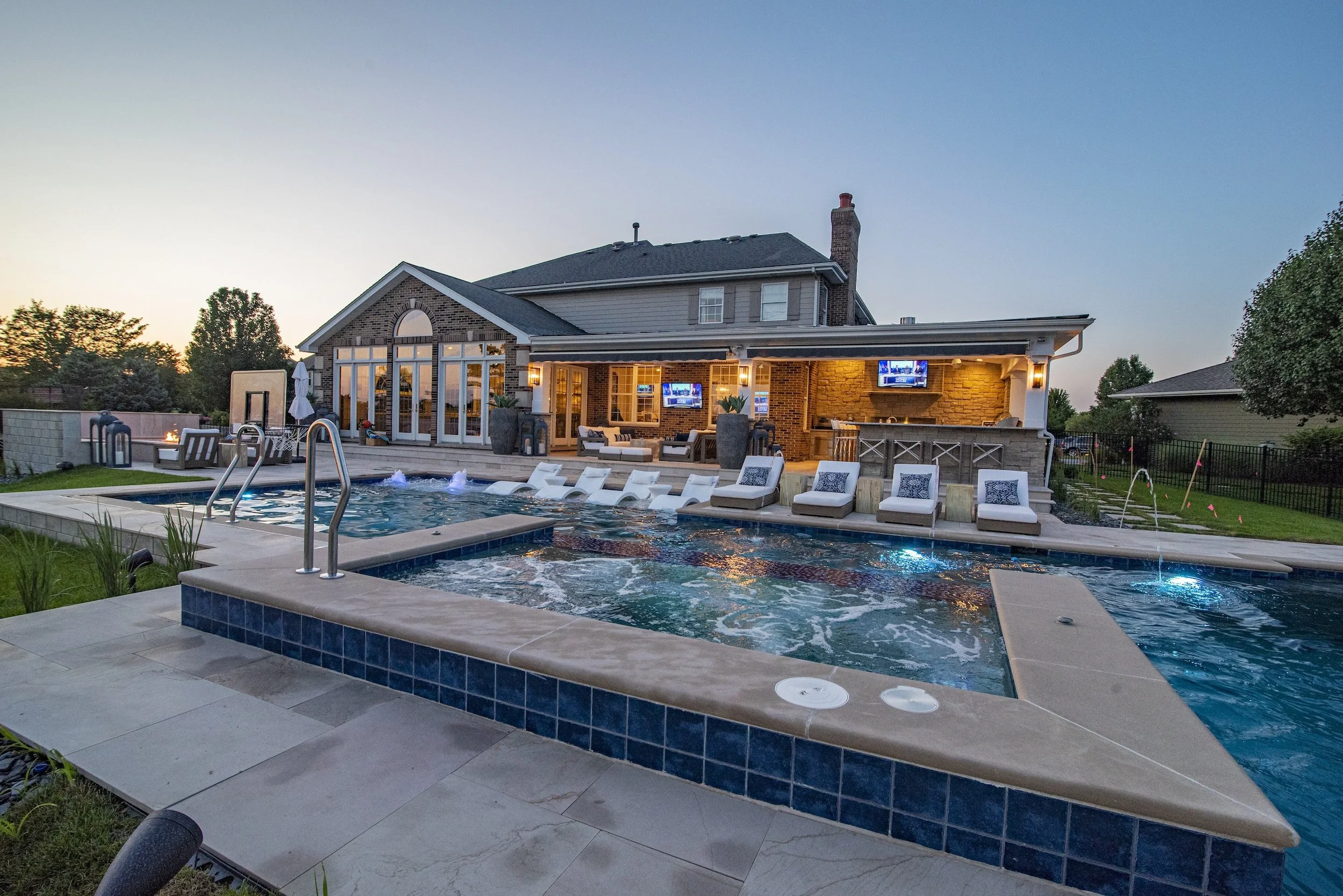 Luxury backyard with a swimming pool, lounge chairs, and a patio connected to a two-story house. The house features large windows, outdoor seating, a bar area, and a fire pit. Trees and lawn surround the pool area, with water fountains in the pool.