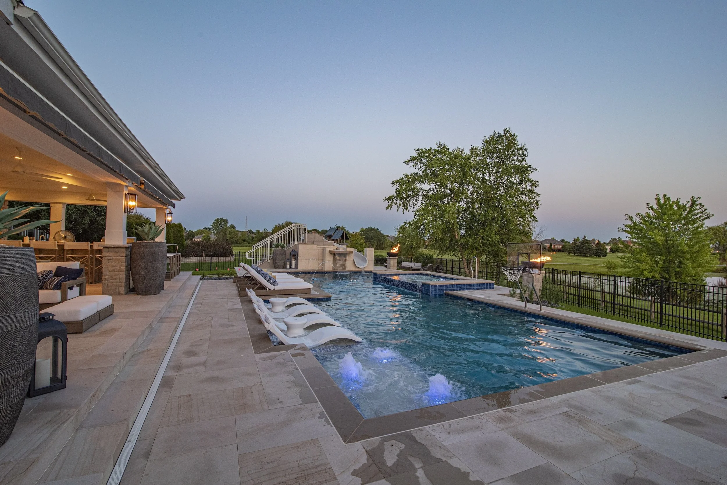 Luxury outdoor patio with swimming pool and water features, surrounded by lounge chairs, oversized planters, and a view of a grassy landscape and trees.