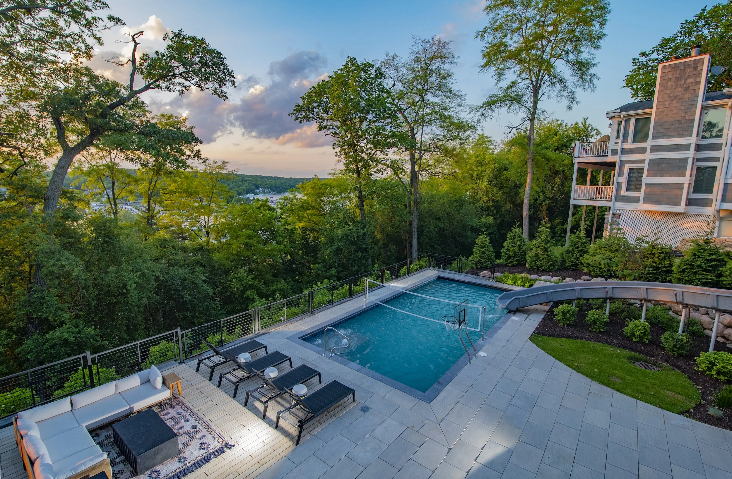 Backyard with a swimming pool, water slide, outdoor seating, and trees surrounding a house.