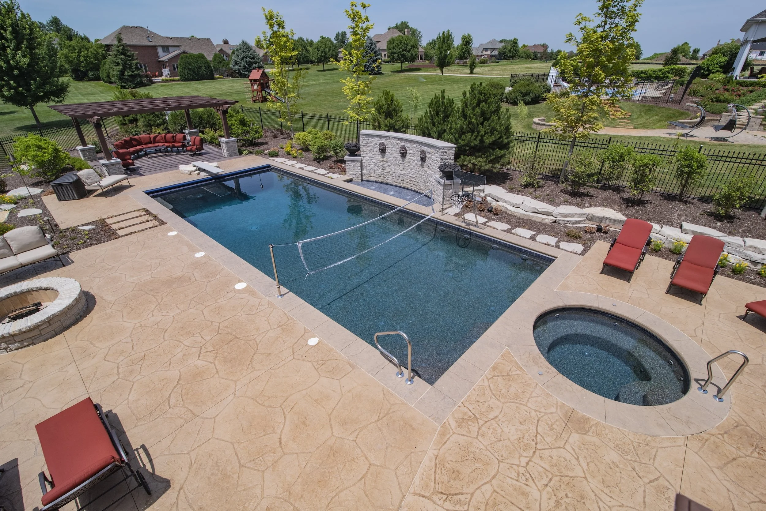A backyard with a swimming pool, hot tub, and outdoor seating area with red lounge chairs. The pool has a volleyball net and water fountain wall. The surrounding landscape includes a grassy lawn, trees, and a fence. There's a fire pit on a stamped co