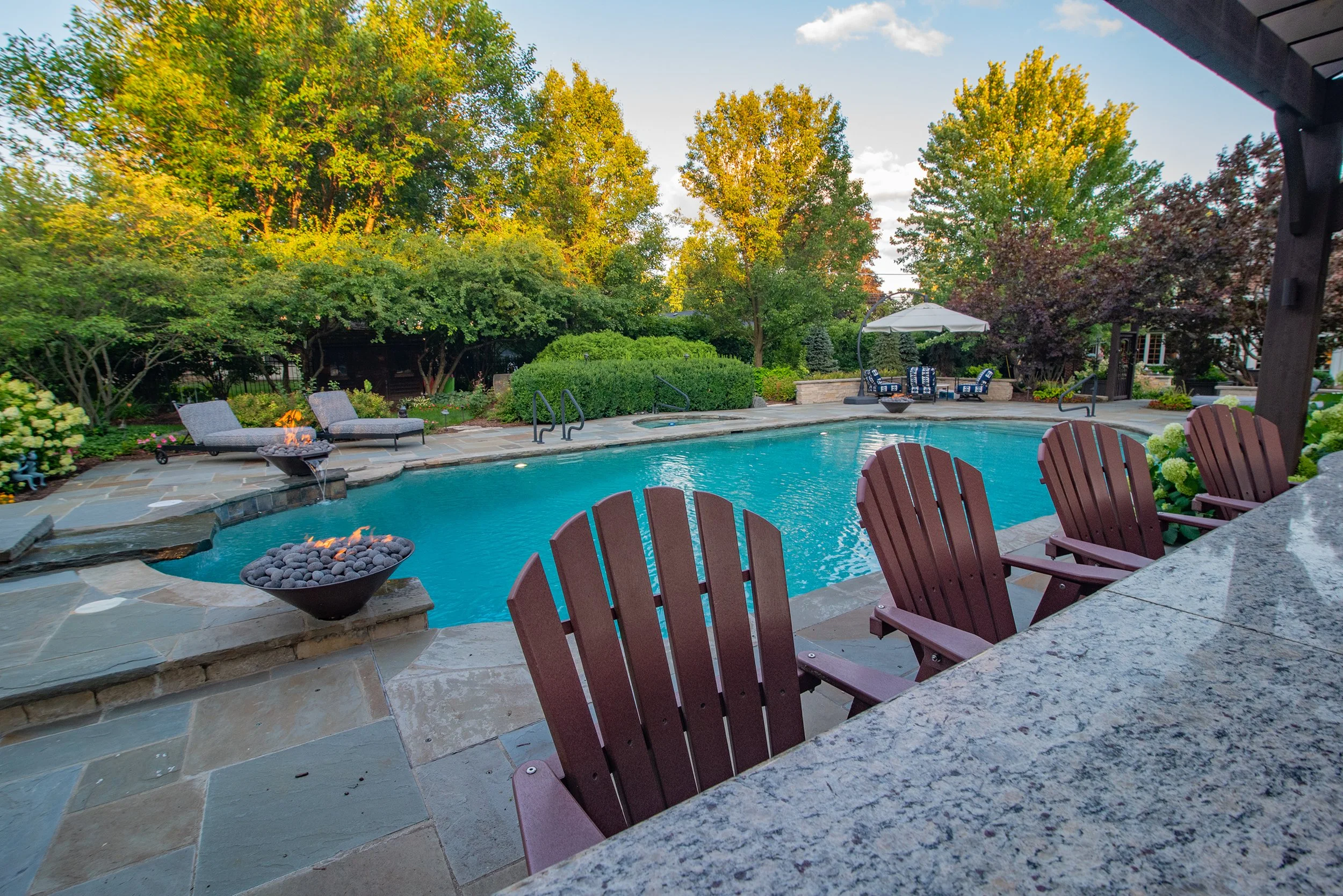 Backyard pool area with wooden chairs, fire features, and lush greenery.