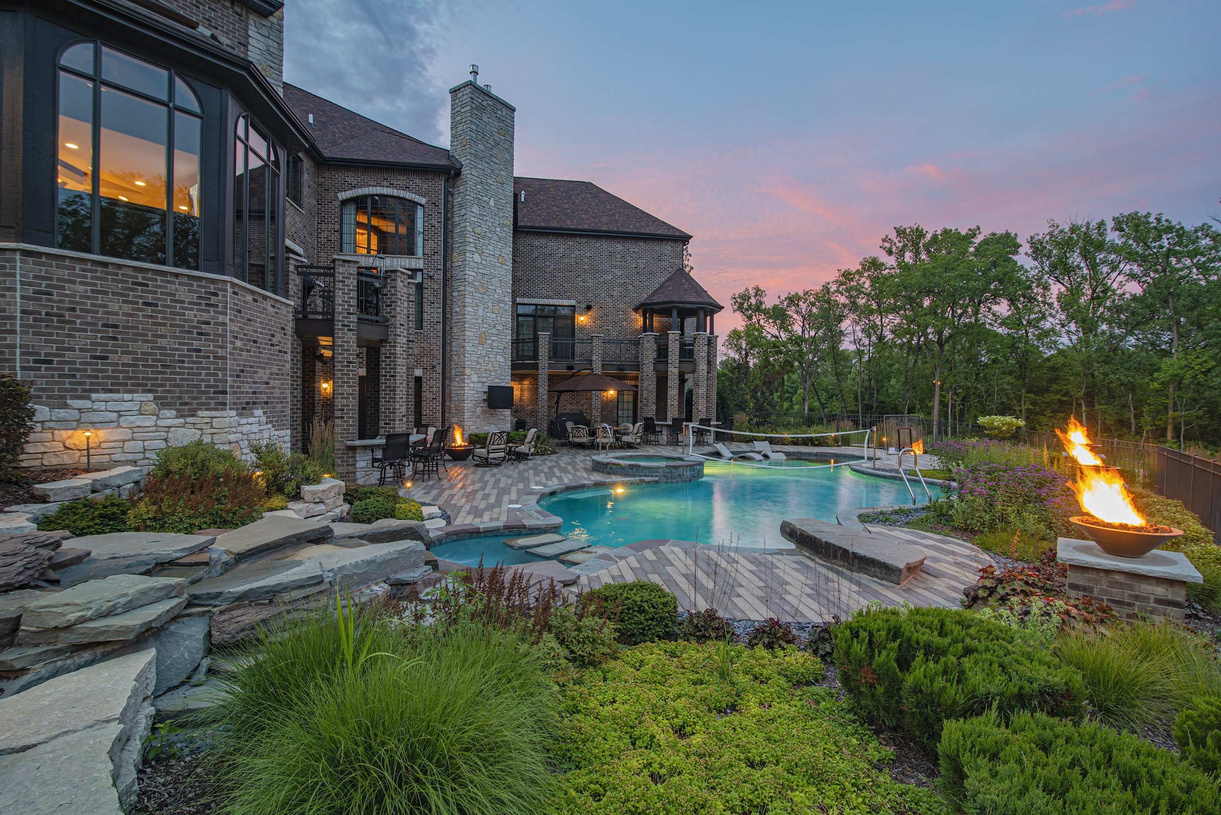 Luxury house exterior with swimming pool, fire pits, patio, manicured gardens, and surrounded by trees at sunset.