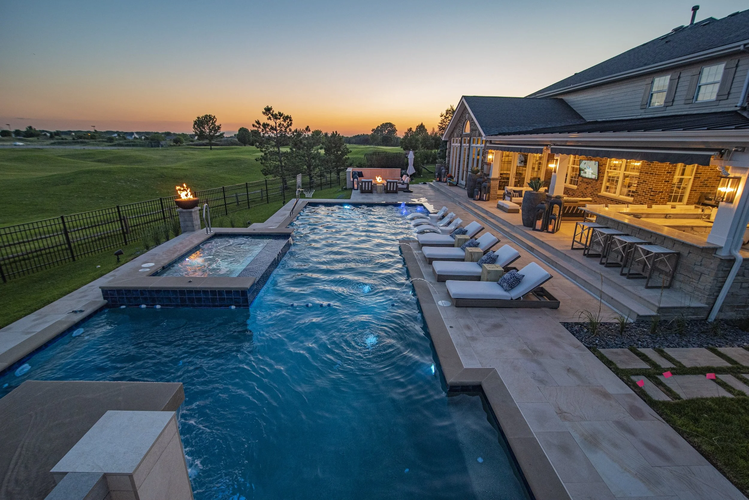 Luxury backyard with a swimming pool and spa, surrounded by lounge chairs and outdoor seating, adjacent to a large house with a patio and outdoor kitchen, set against a sunset backdrop.