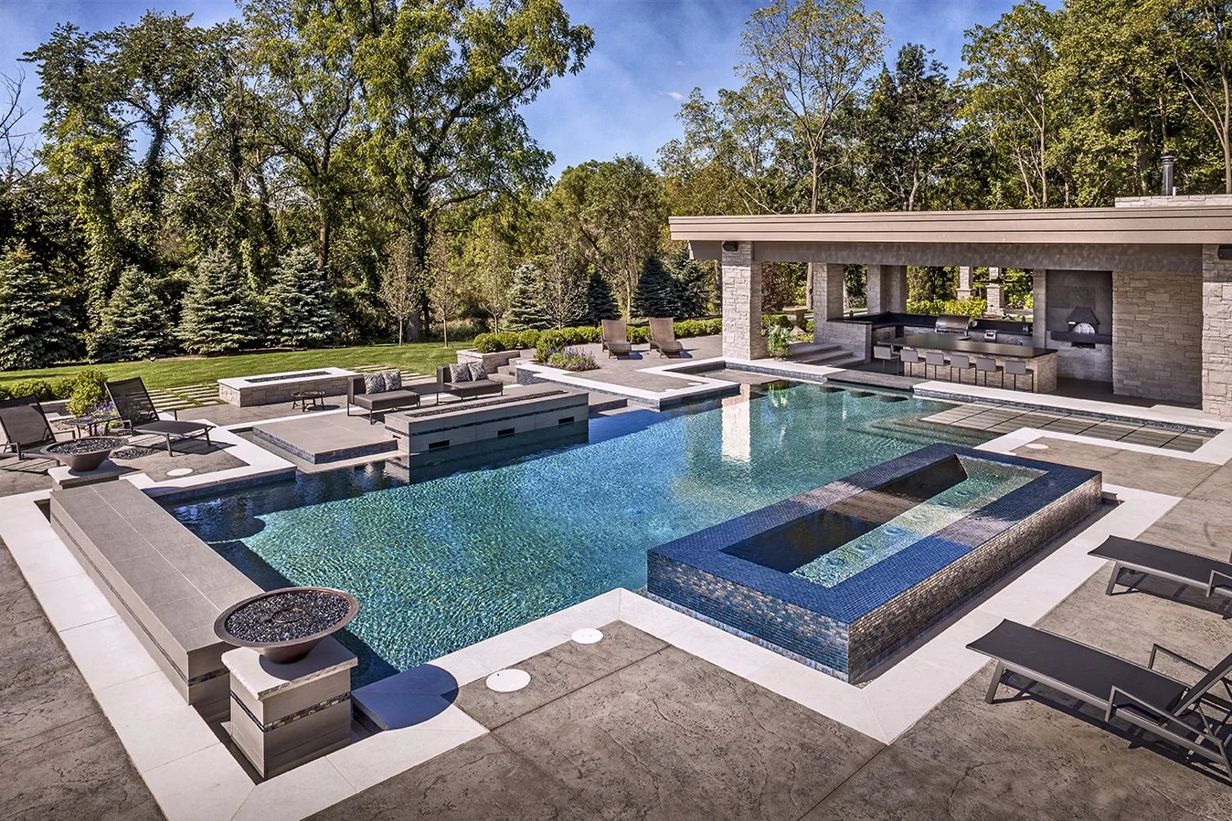 Modern backyard with a large swimming pool, sun loungers, and an outdoor kitchen area. The setting includes trees and neatly trimmed grass, creating a luxurious outdoor living space.