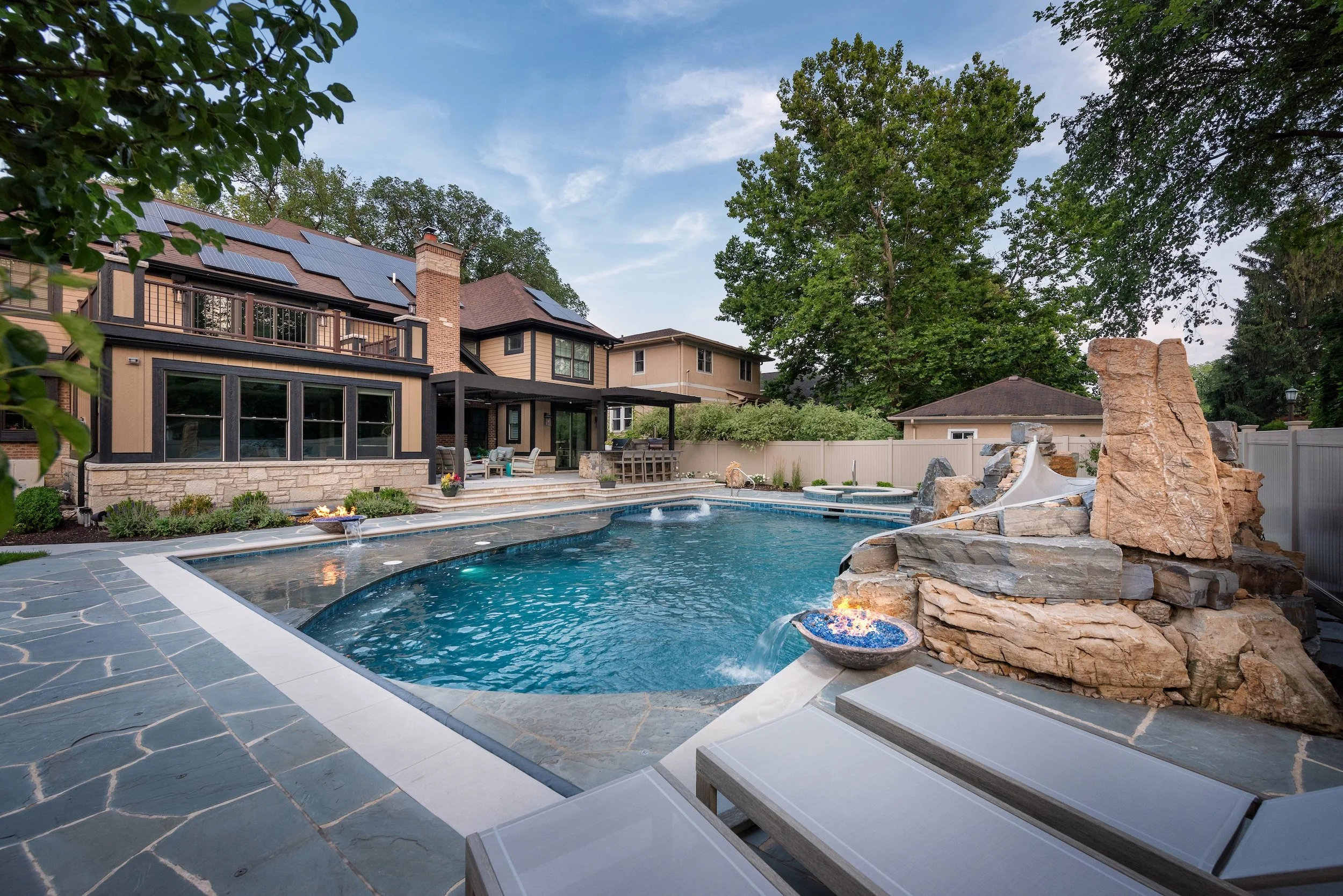 Luxurious backyard featuring a modern swimming pool surrounded by a stone patio, fire pit, and outdoor seating area. A large, two-story house with solar panels is in the background. Lush greenery and trees encircle the perimeter.