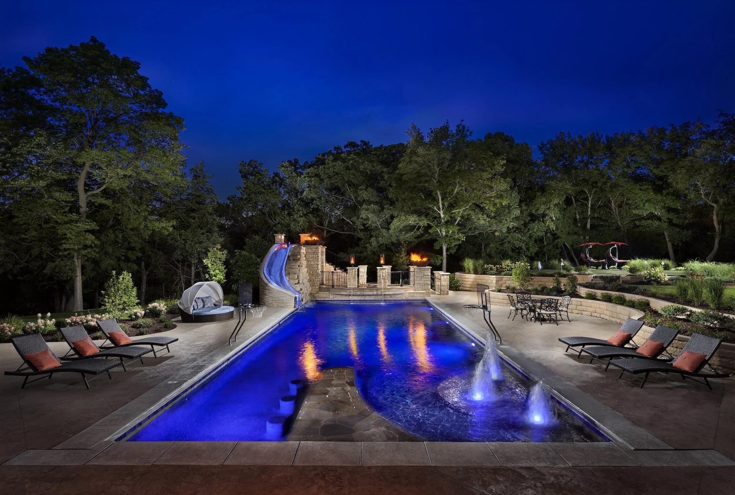Luxurious backyard with illuminated swimming pool, water slide, lounge chairs, and lush surrounding trees at night.