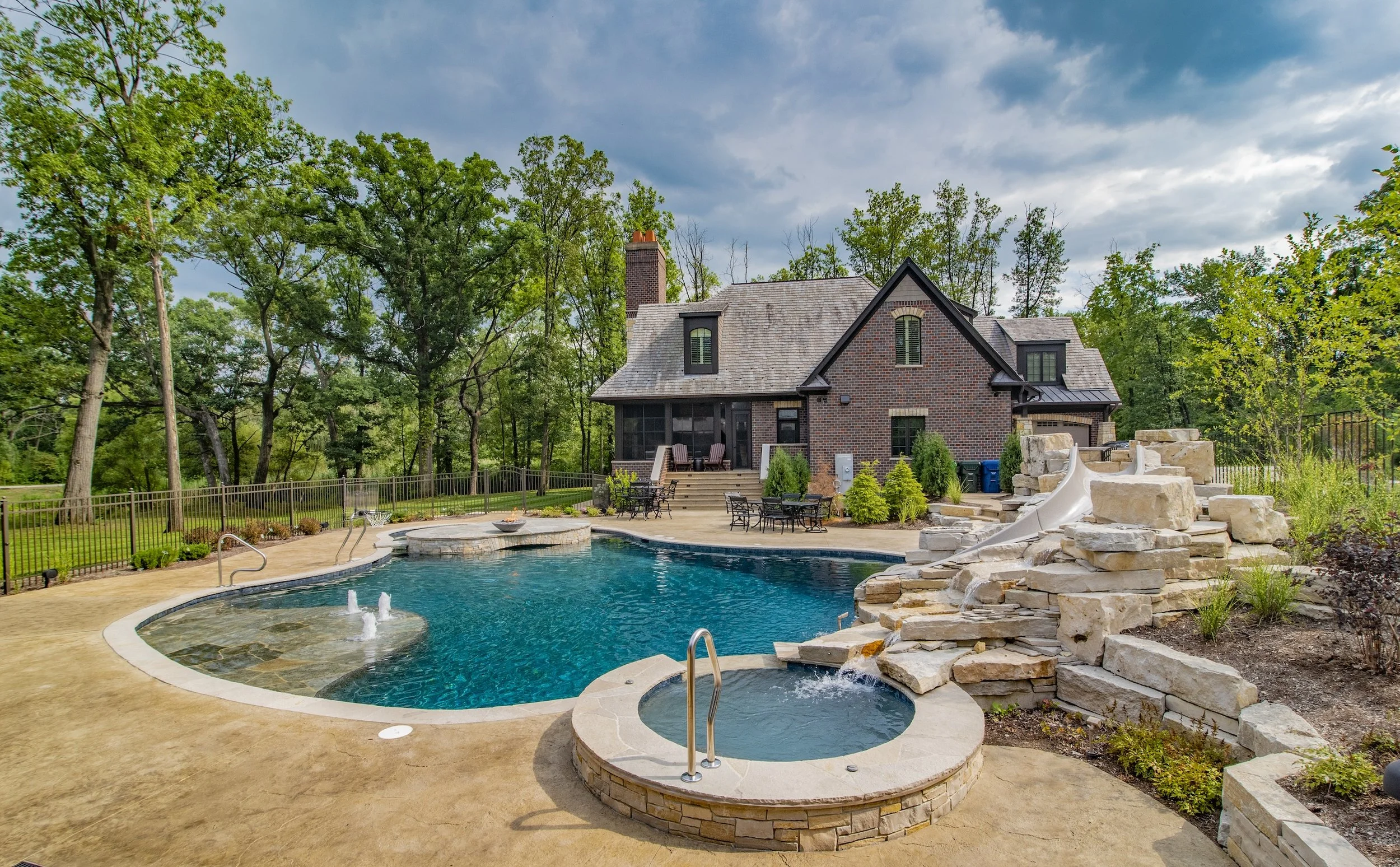 Residential backyard with a swimming pool, hot tub, and rock slide next to a brick house surrounded by trees.