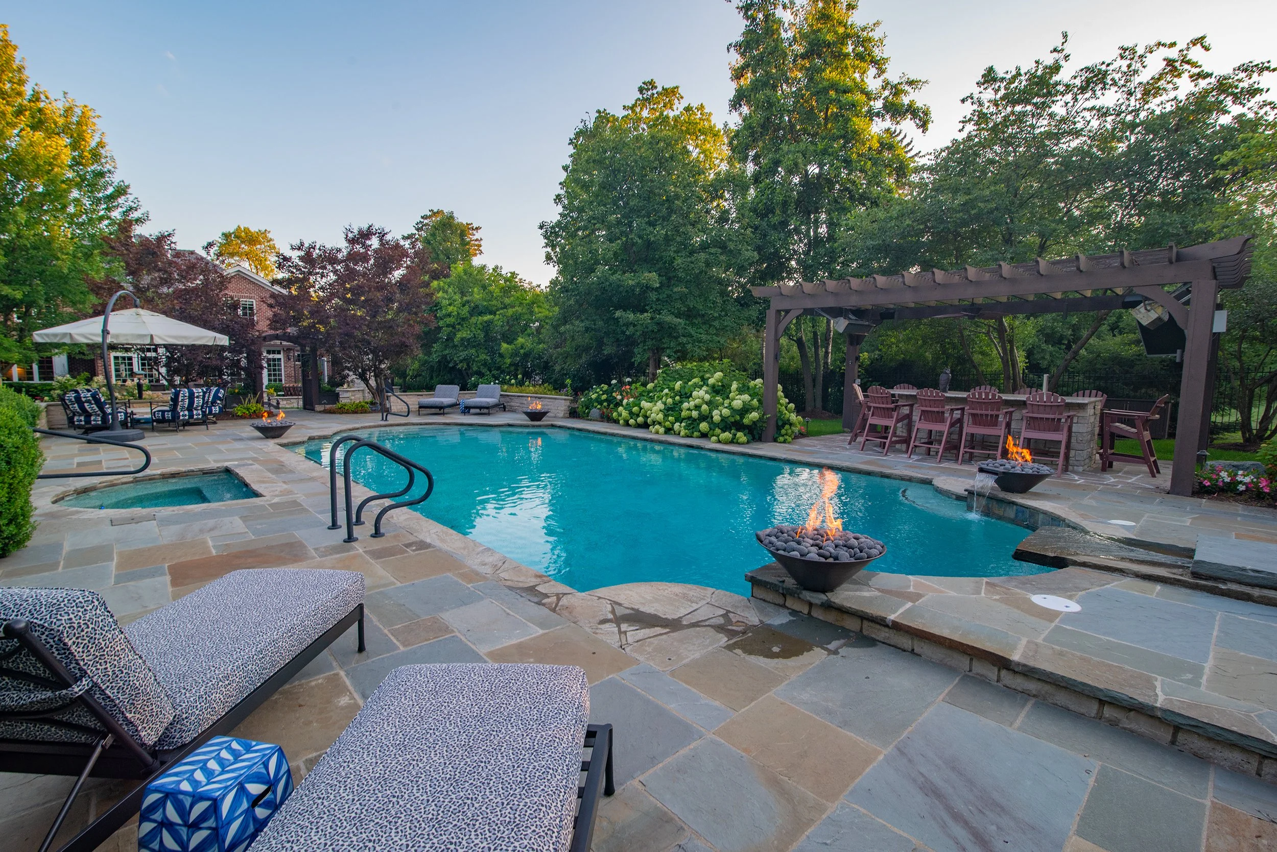 Backyard with a swimming pool, lounge chairs, hot tub, and a pergola with seating, surrounded by trees and plants.