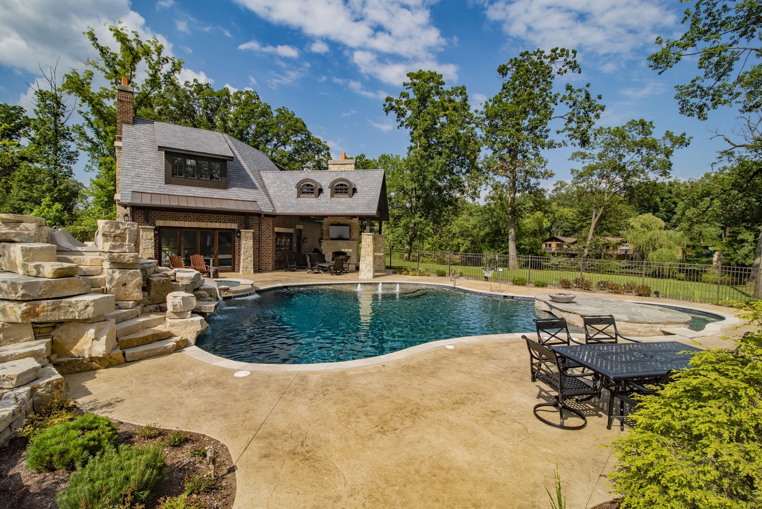 Luxury backyard with pool, spa, patio furniture, and brick house surrounded by trees.