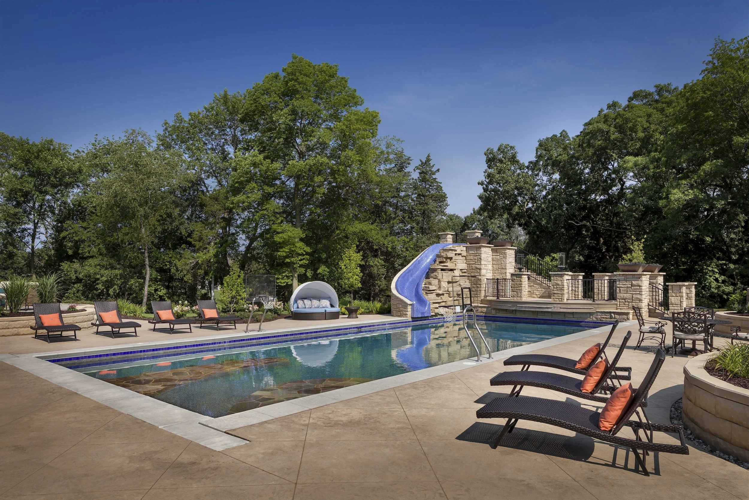 Luxury backyard pool area with a water slide, lounge chairs, canopy bed, patio furniture, stone patio, and surrounding trees and plants.