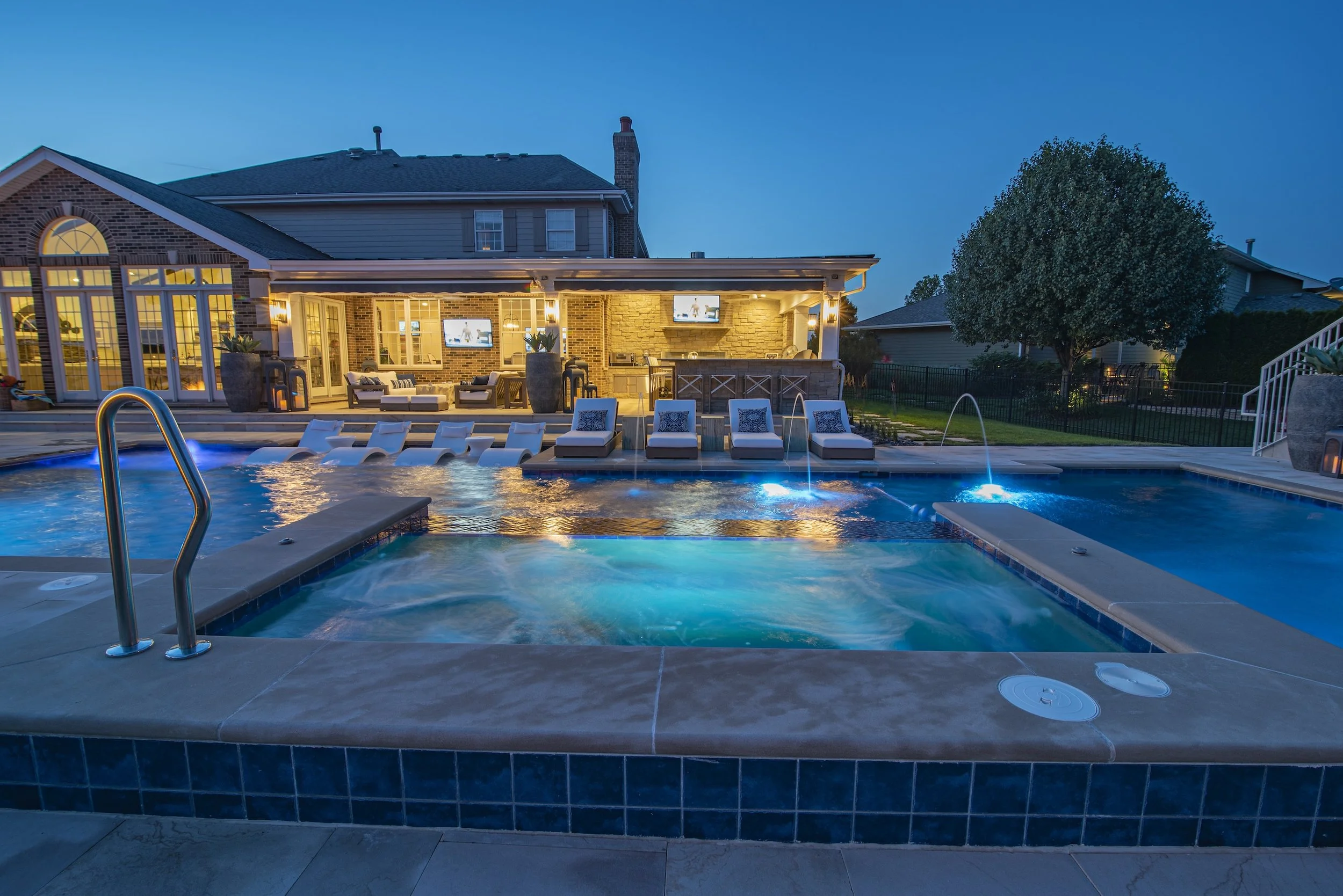 A modern backyard with a pool and patio at dusk, featuring lounge chairs, a hot tub, outdoor seating, and a well-lit house.