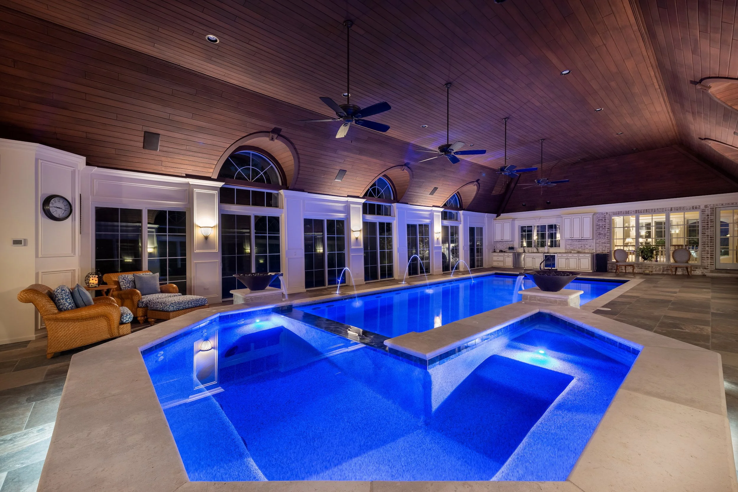 Indoor swimming pool with blue lighting, surrounded by luxury seating, high wooden ceiling with ceiling fans, and multiple windows for natural light.