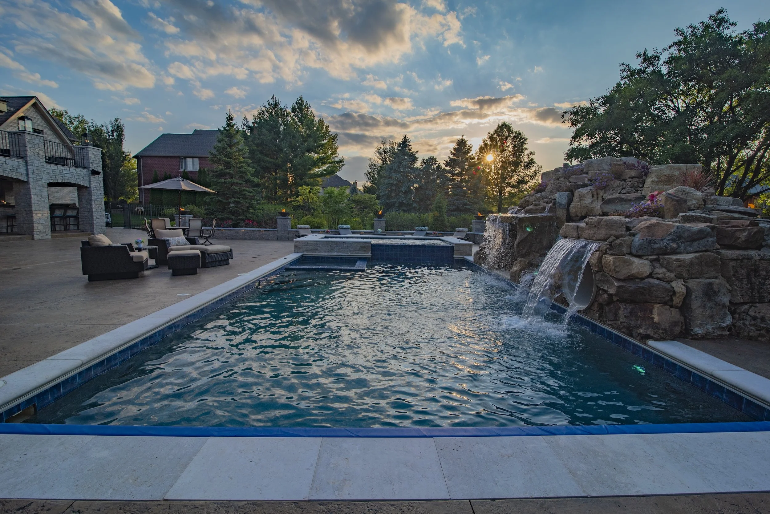 Outdoor swimming pool with rock waterfall feature, surrounded by patio furniture and trees at sunset.