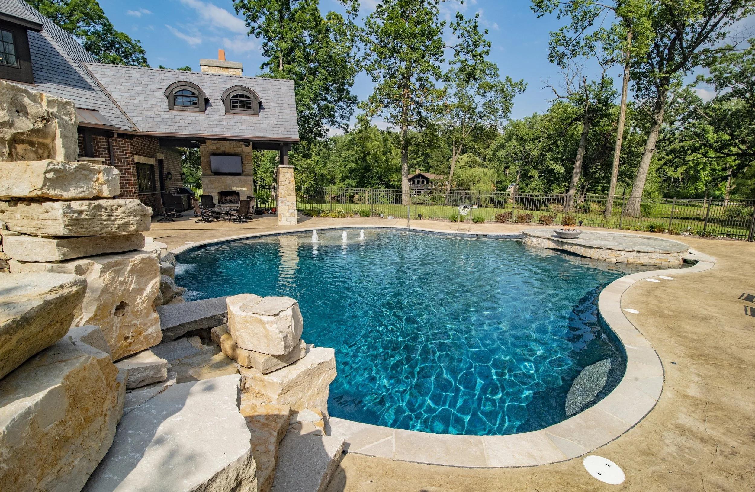 Outdoor pool with stone patio, rock waterfall, seating area, and surrounding trees.