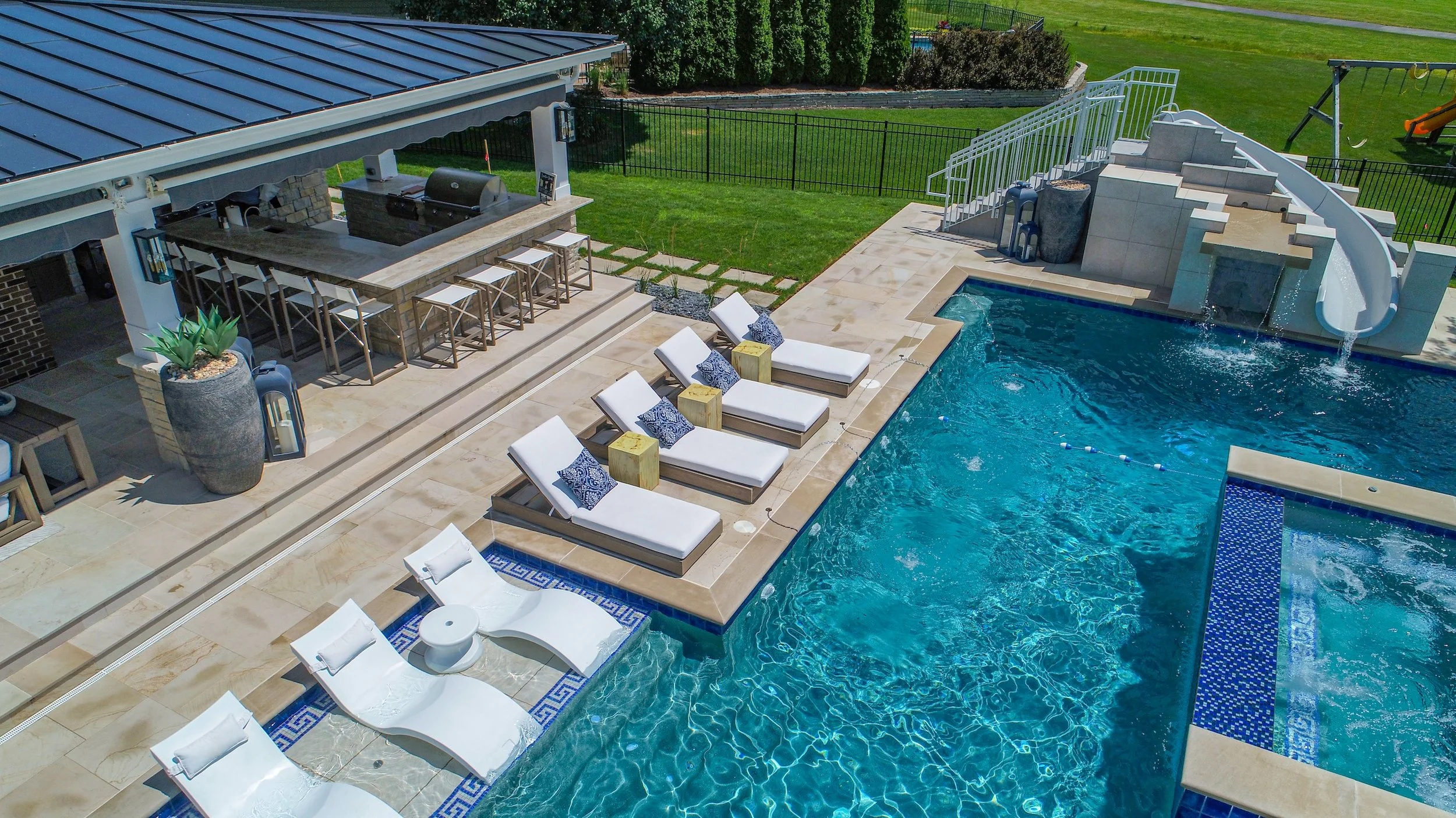 Outdoor pool area with lounge chairs, a water slide, poolside bar with stools, and a covered patio with a grill.