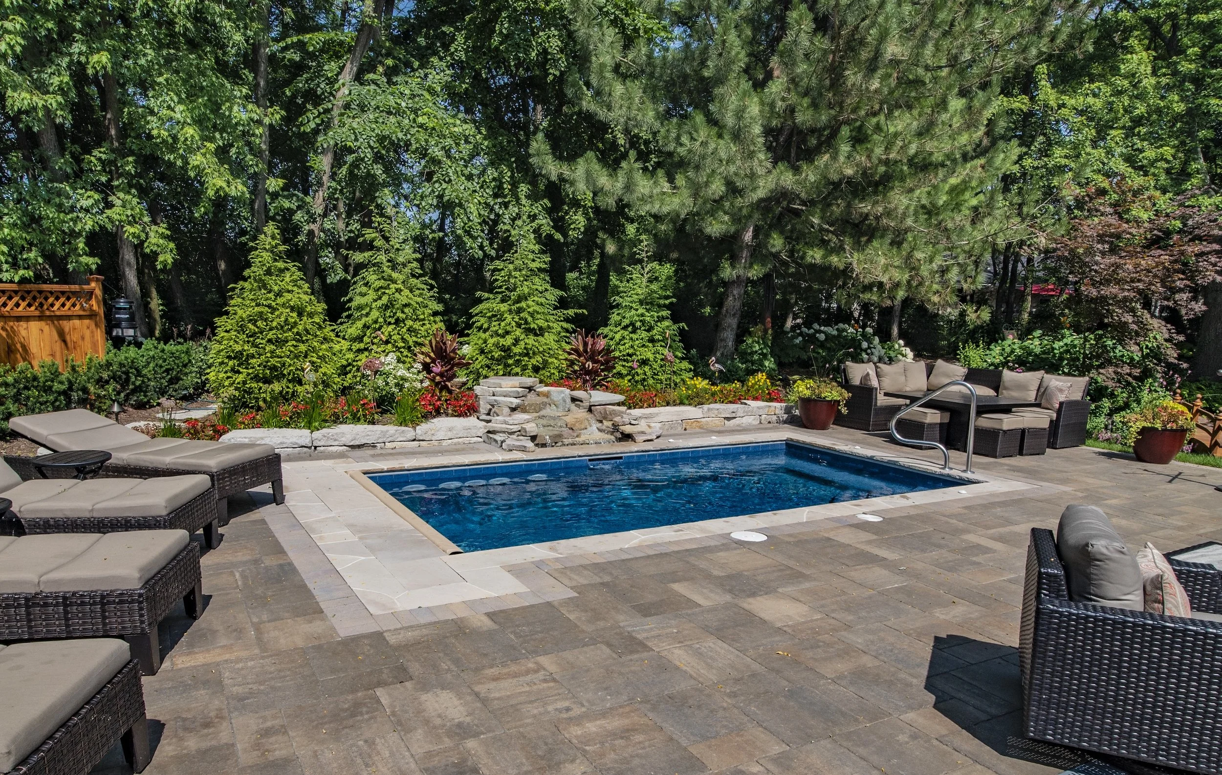 Outdoor patio with a small swimming pool, surrounded by lounge chairs, wicker furniture, and decorative plants, with a backdrop of tall trees.