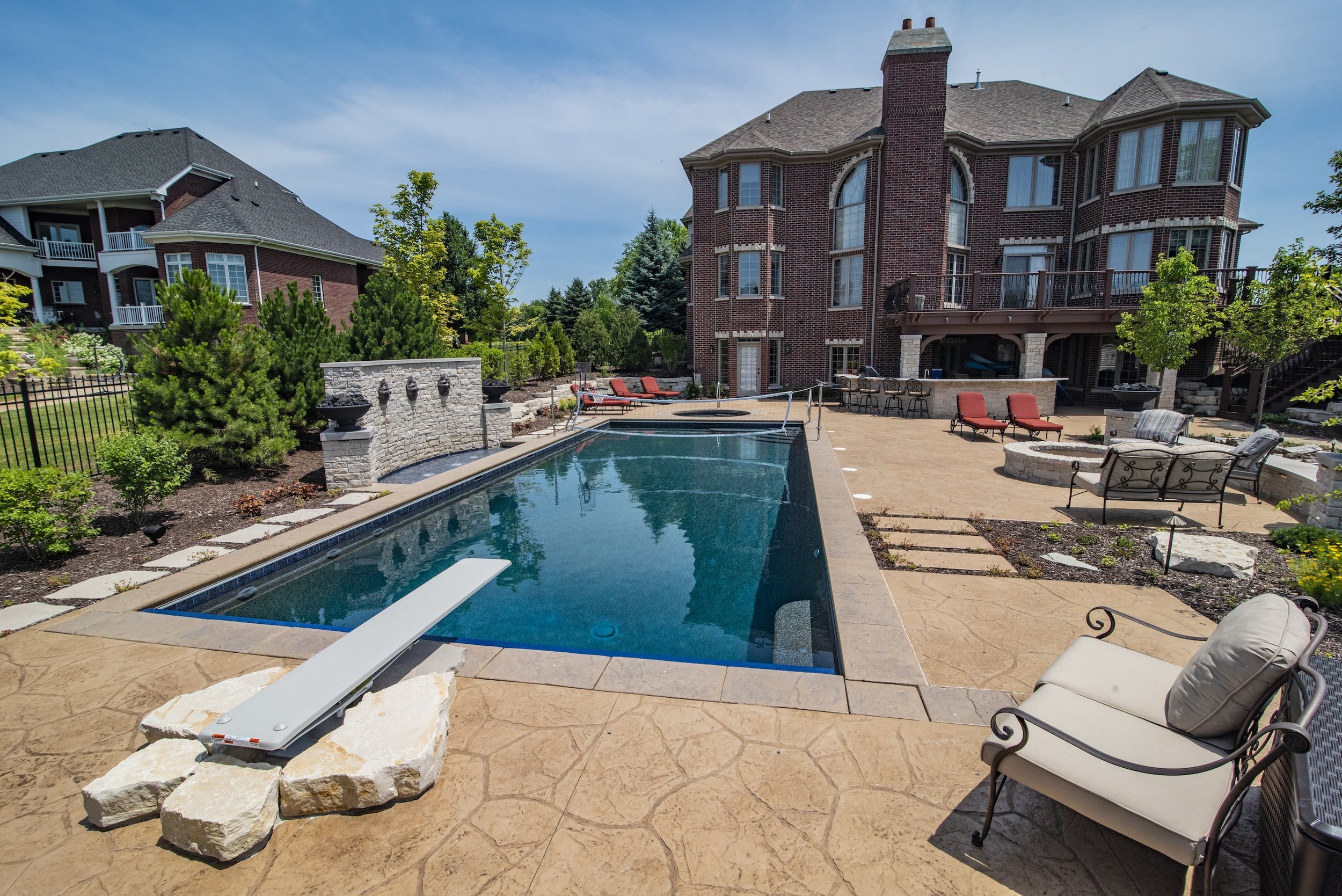 Backyard of a large brick house with a rectangular swimming pool, diving board, patio area, seating, and landscaping.