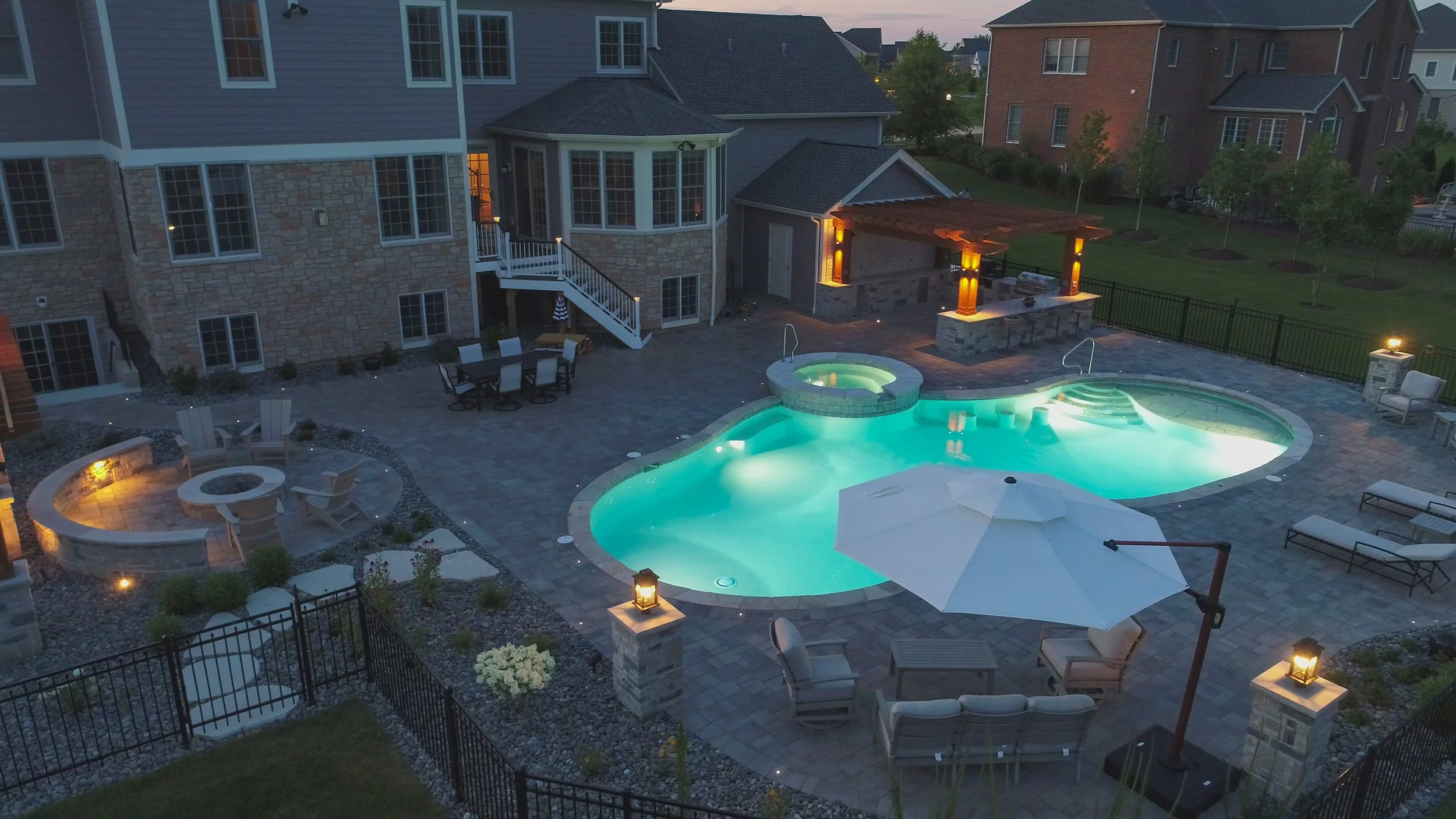 Backyard with illuminated swimming pool, hot tub, patio furniture, fire pit, outdoor kitchen, and surrounding landscaping at dusk.