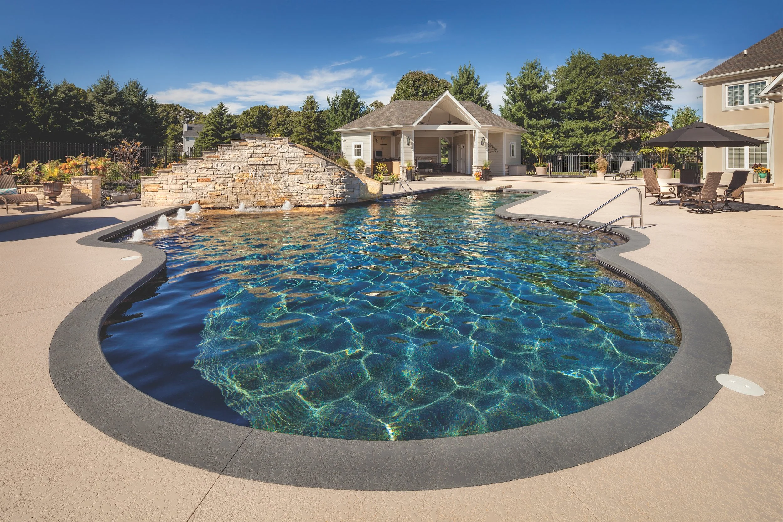 Backyard with a kidney-shaped swimming pool, stone waterfall, poolside furniture, beige patio, and a small pool house with trees in the background.