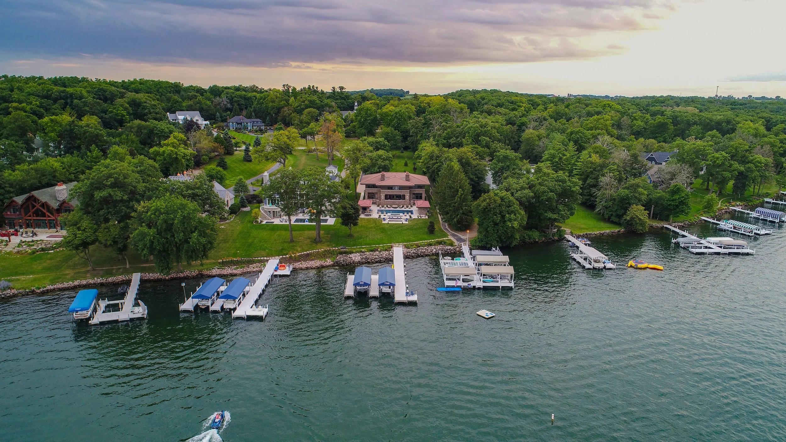 Aerial view of a lakeside mansion surrounded by greenery, with multiple docks and boats on the water.
