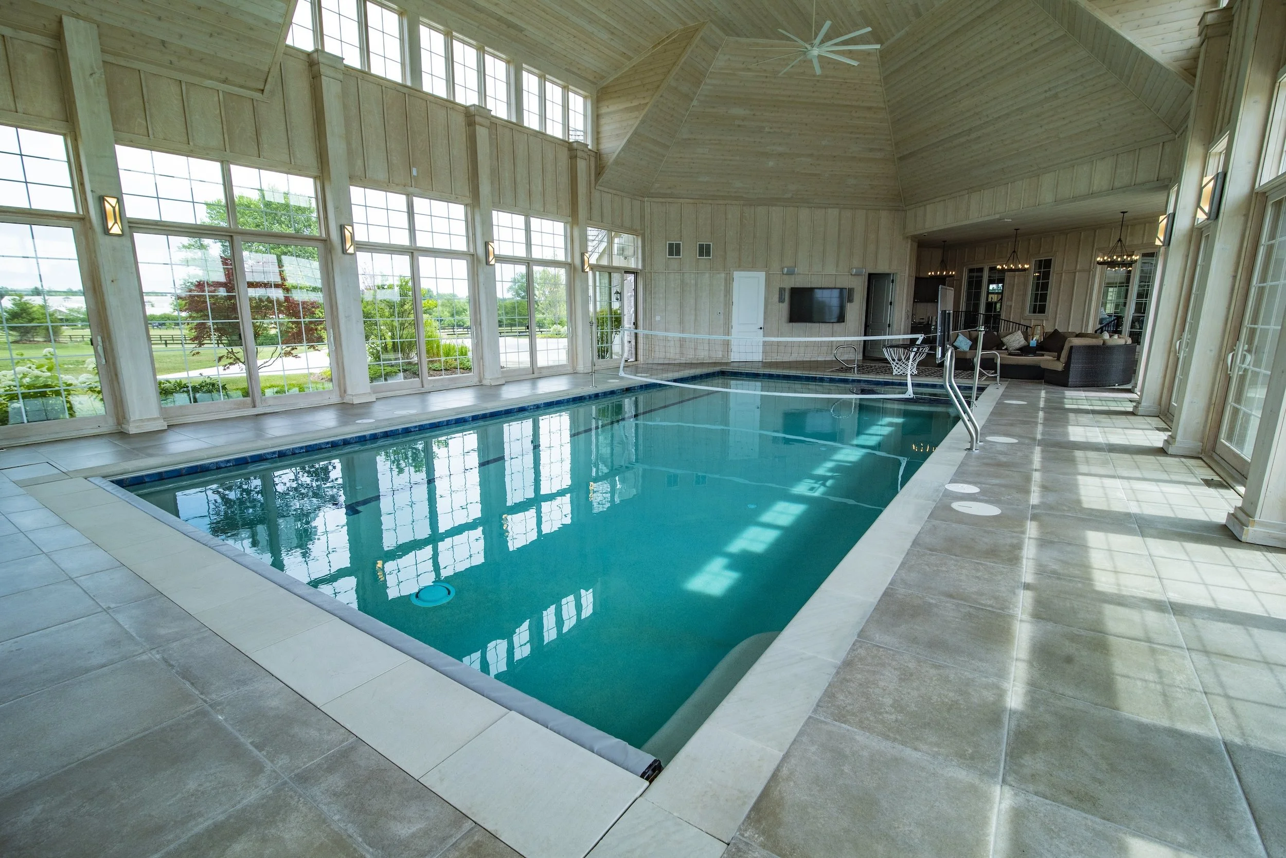 Indoor swimming pool with tall windows and a vaulted wooden ceiling, featuring natural light and outdoor views, with a seating area and a small basketball hoop.