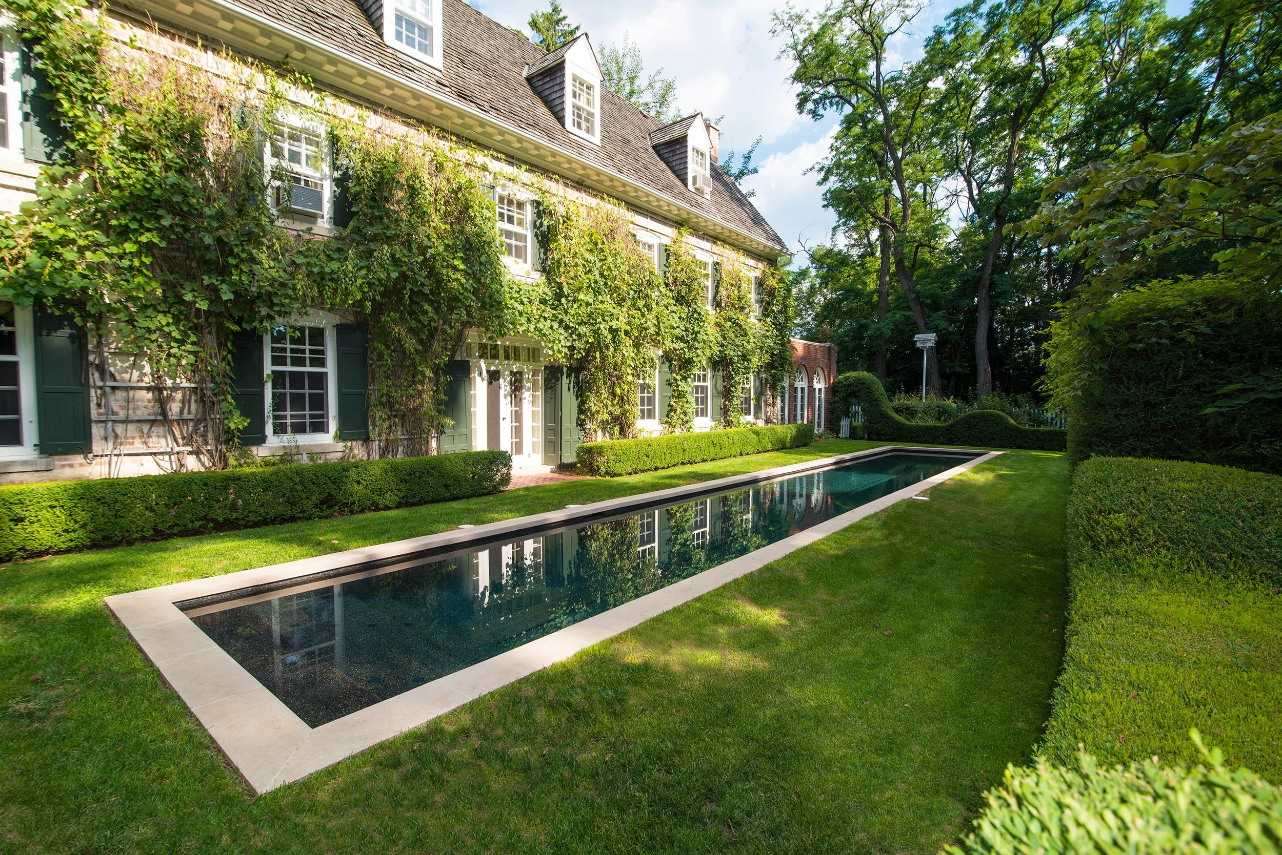 Elegant mansion with ivy-covered facade, long rectangular pool, manicured lawn, and lush greenery.