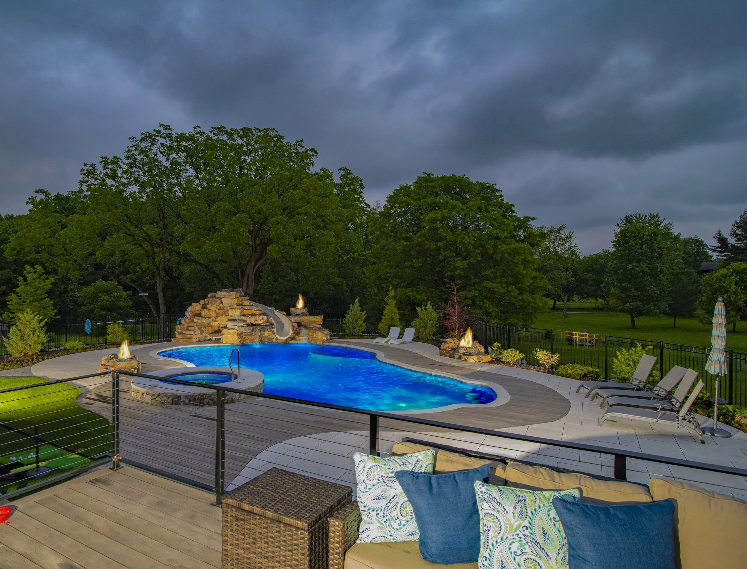 Outdoor pool area with waterfall and fire features at dusk, surrounded by lounge chairs and trees in the background.