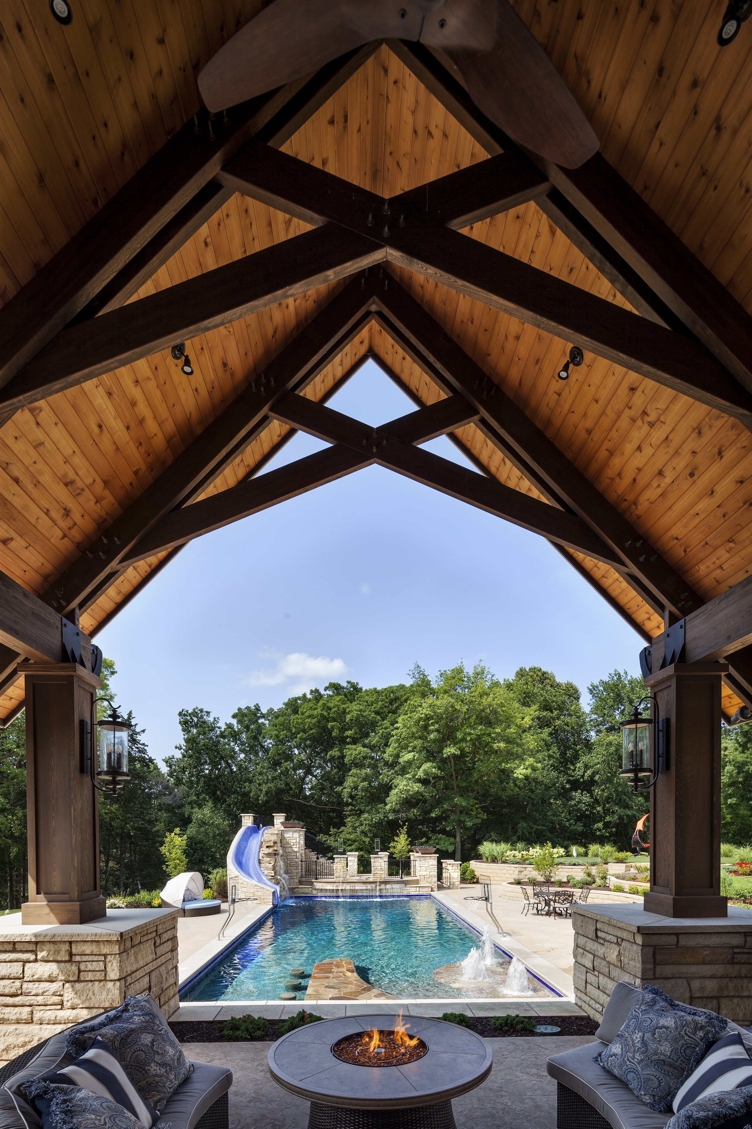 View of a luxurious backyard pool area with a wooden pavilion, stone fireplace, and lush greenery. The scene features a swimming pool with a water slide, surrounded by a landscaped garden.