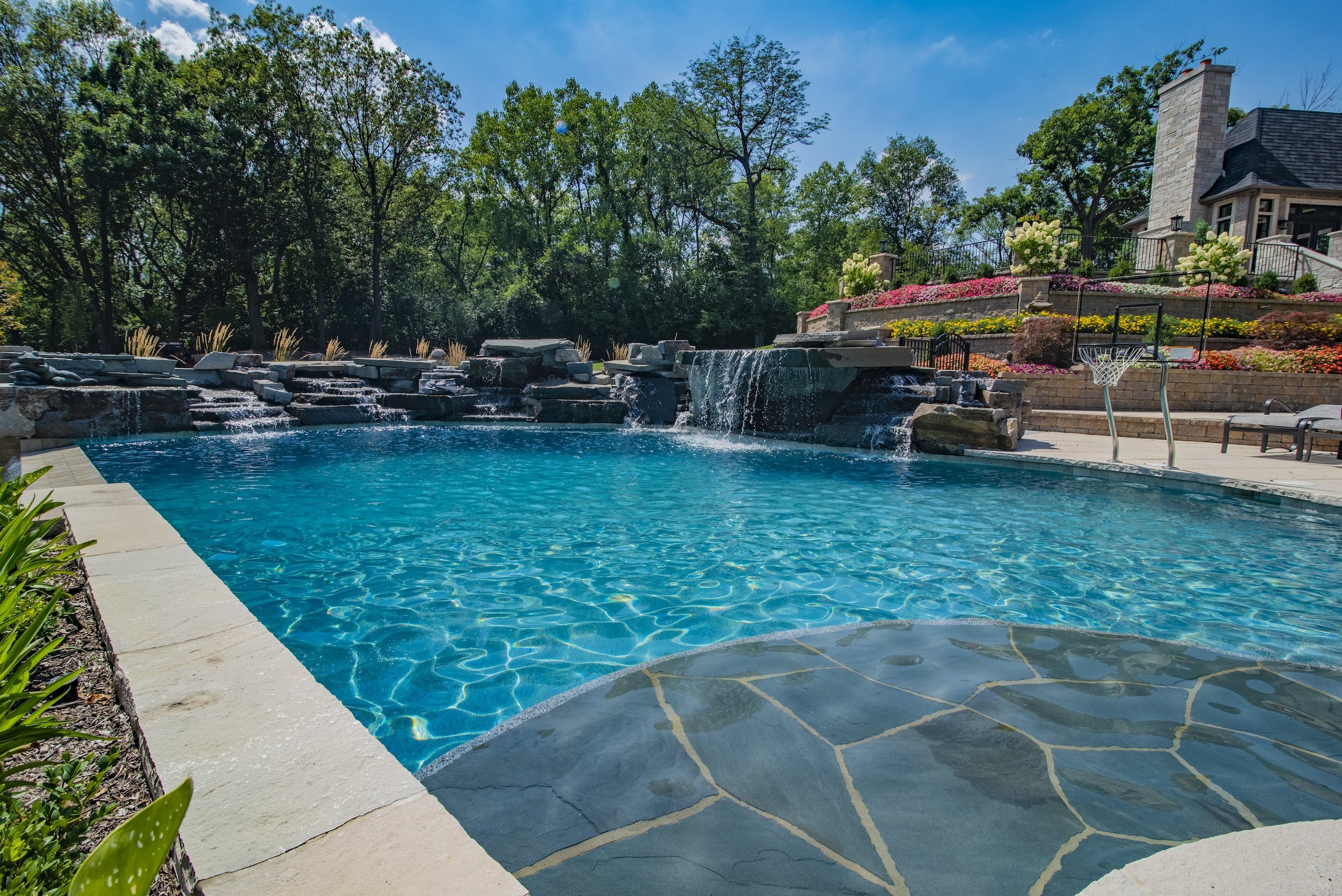 Luxury swimming pool with waterfall feature, surrounded by landscaped gardens and a house in the background.