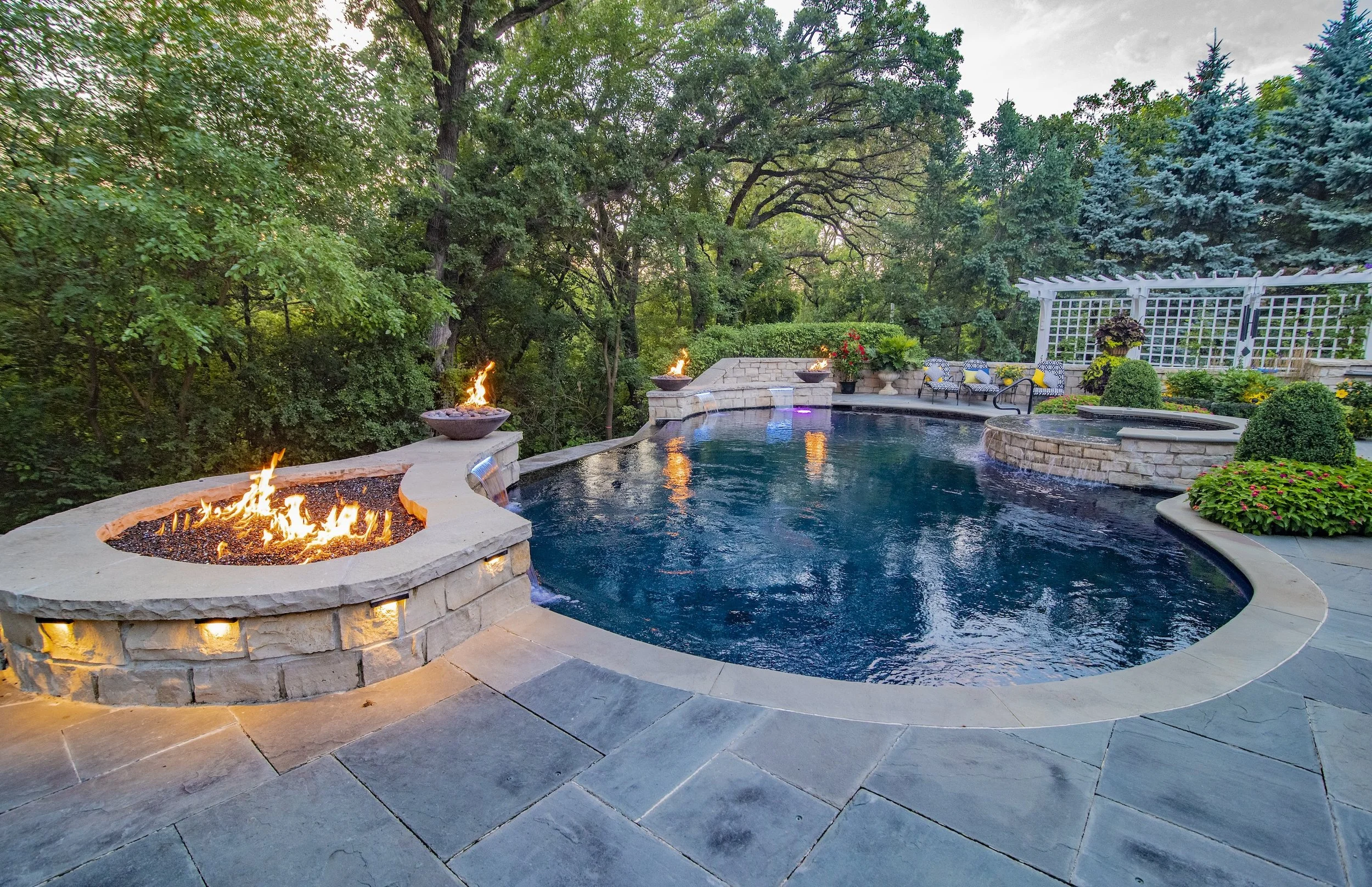 Luxurious backyard with a curved pool, stone patio, fire pits, and lush greenery.