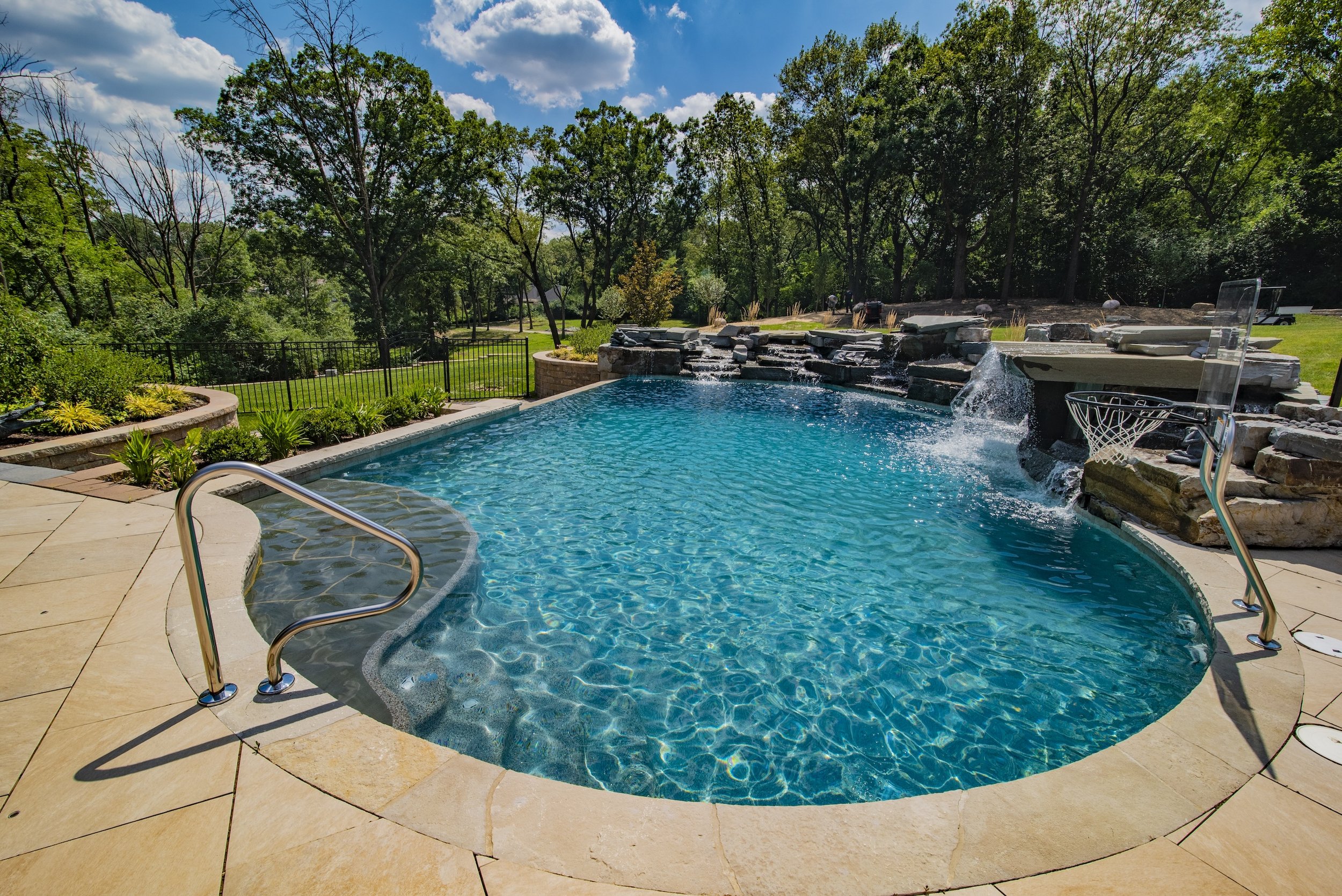 Backyard swimming pool with waterfall feature, surrounded by stone tiles and landscaping, under a sunny sky.
