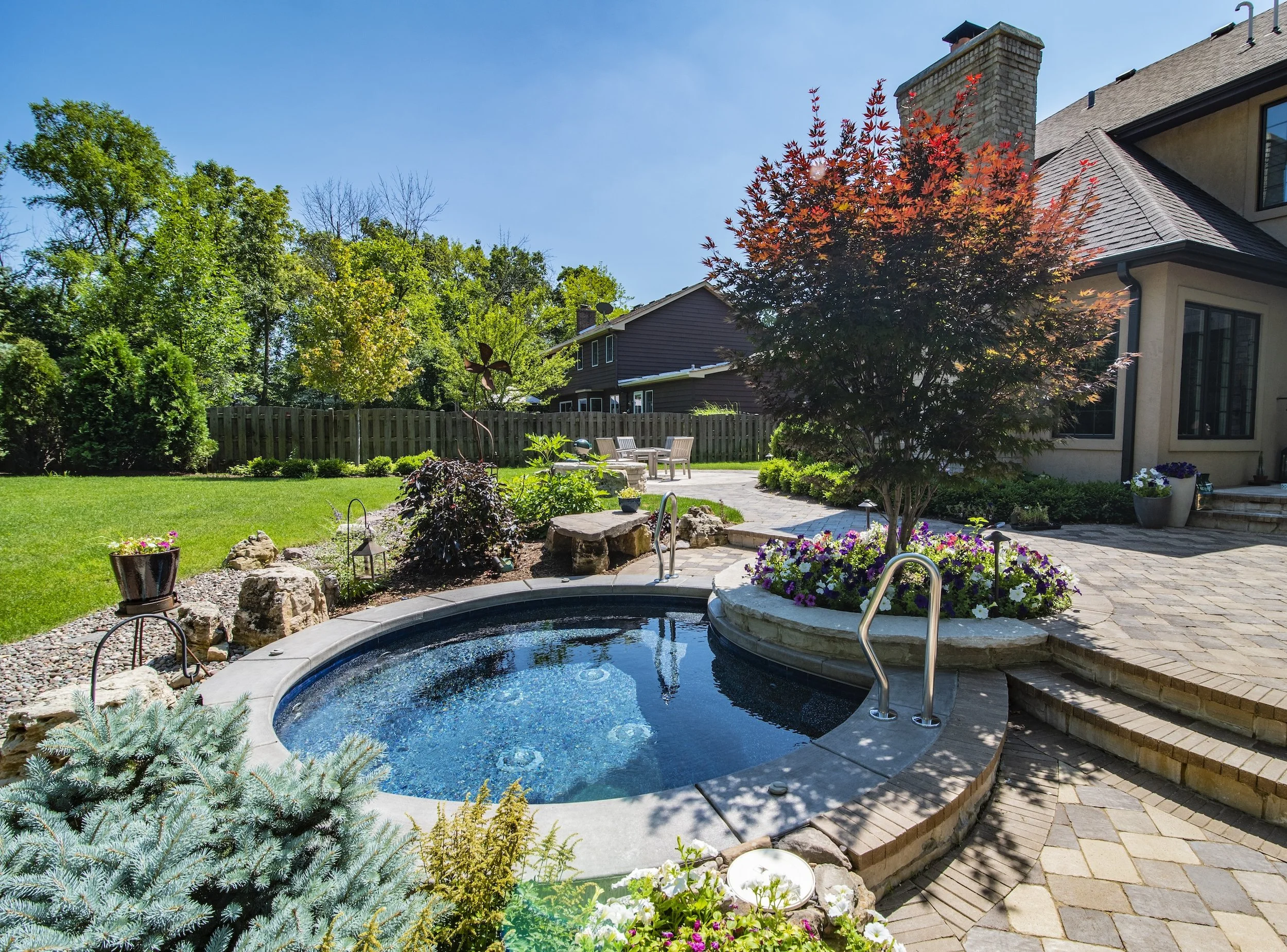 Backyard with a small circular pool, lush garden, stone patio, and outdoor seating area under sunshine.