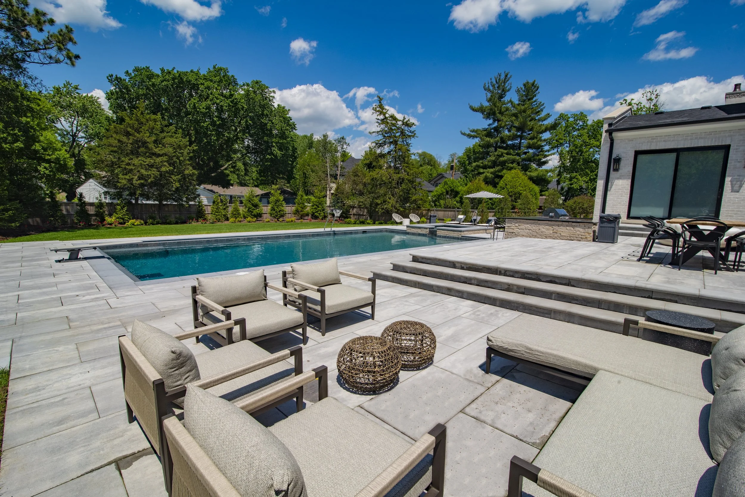 Outdoor patio area with a swimming pool, lounge chairs, and a table set. Surrounded by trees and a clear blue sky.