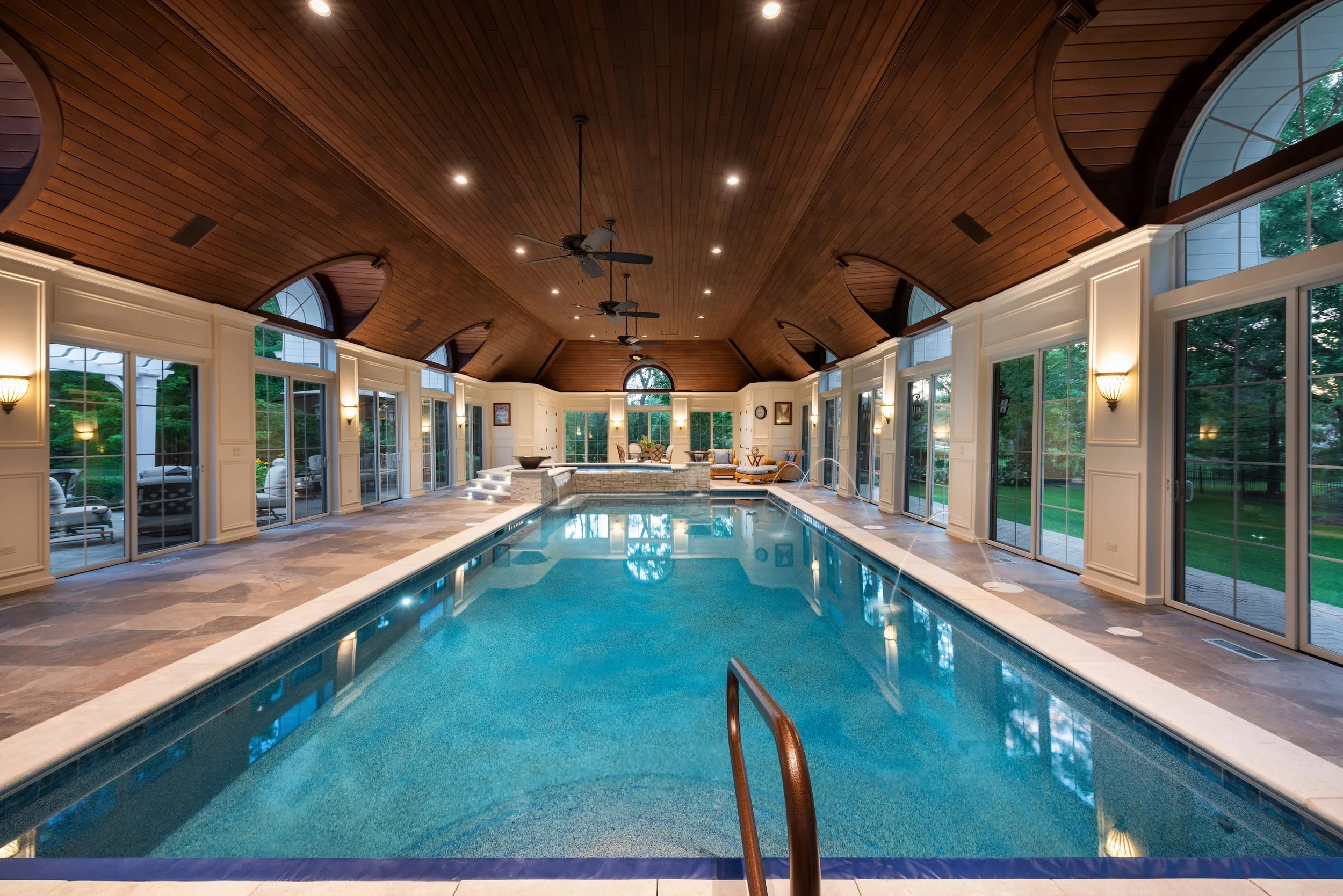 Indoor swimming pool with a wooden vaulted ceiling, large windows, and ambient lighting. The pool area includes lounging furniture and decorative elements.