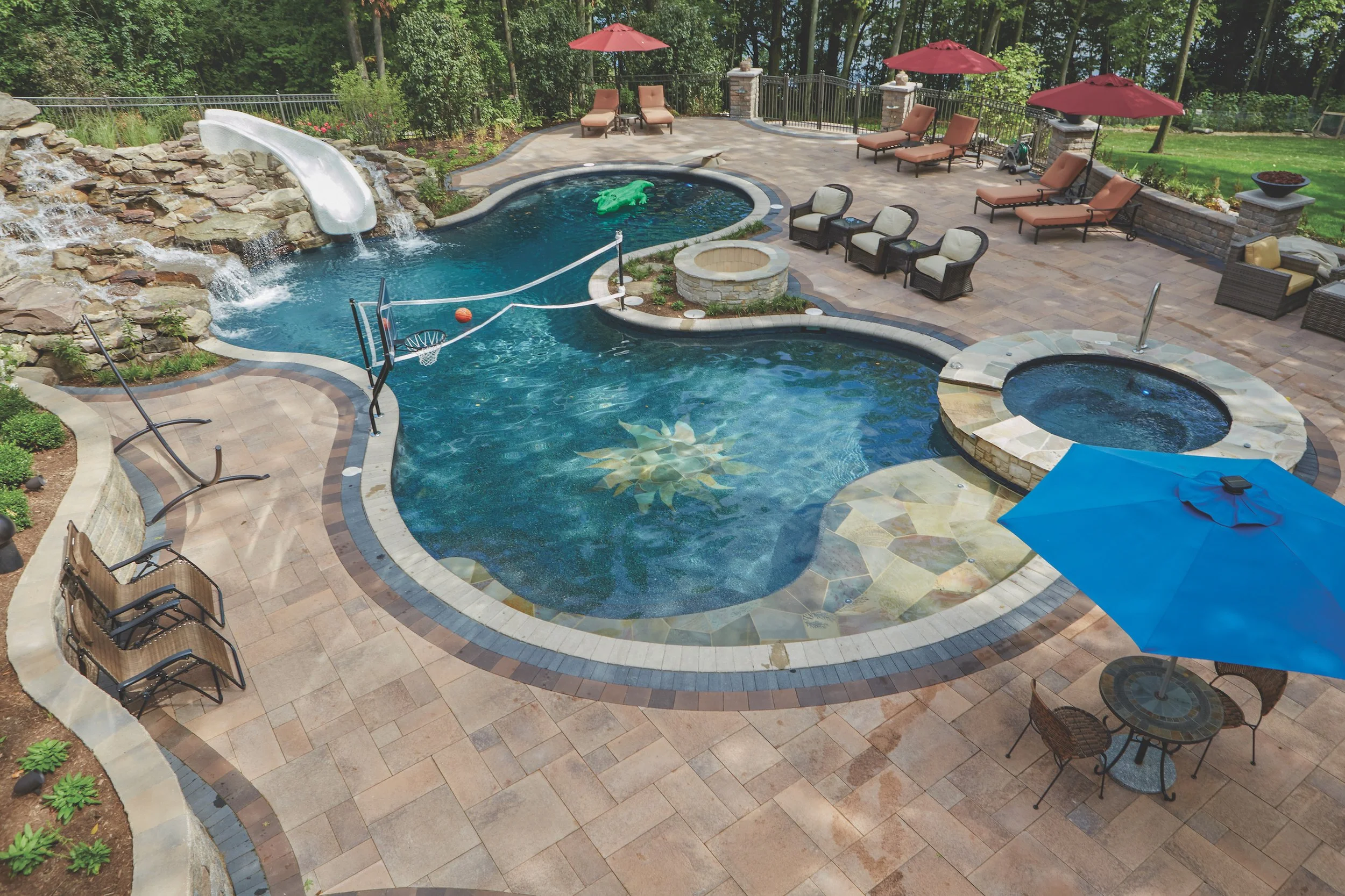 A luxurious backyard pool area featuring a large swimming pool with a decorative design, a water slide, and a spa. The pool deck has several lounge chairs, red umbrellas, and a blue umbrella over a table with chairs. A volleyball net spans a section 