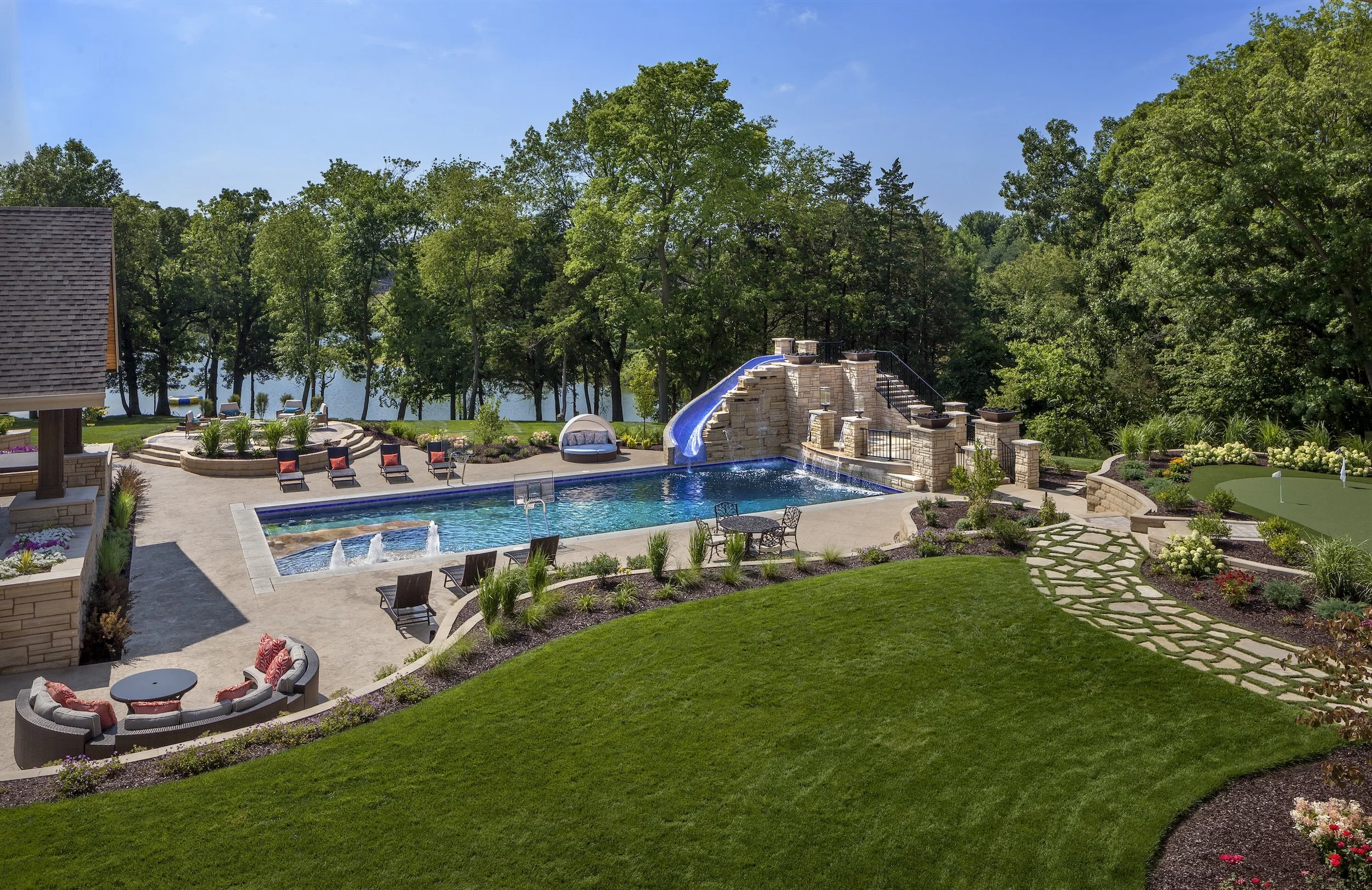 Luxurious backyard with a swimming pool, lounge chairs, water slide, and landscaped garden, surrounded by trees.