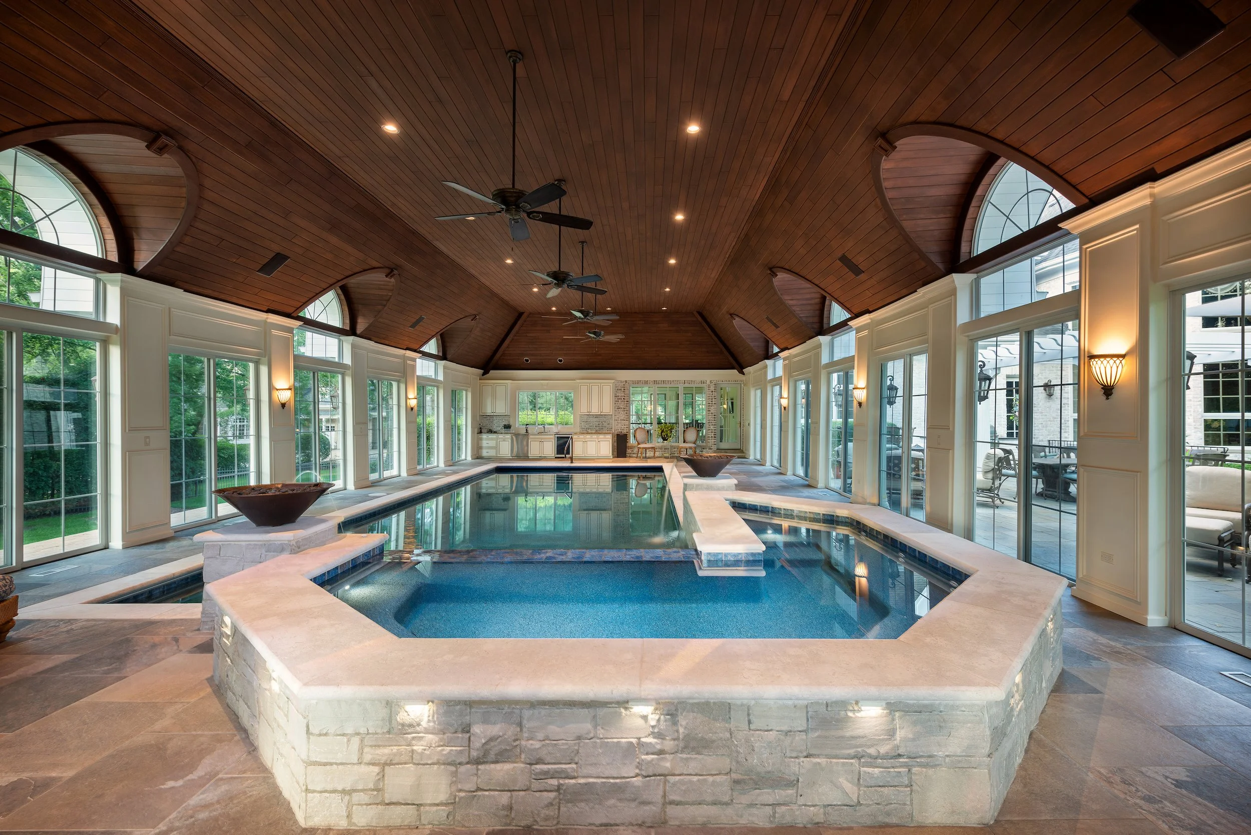 Luxurious indoor swimming pool with wooden ceiling, ceiling fans, large windows, stone accents, and a spacious seating area.
