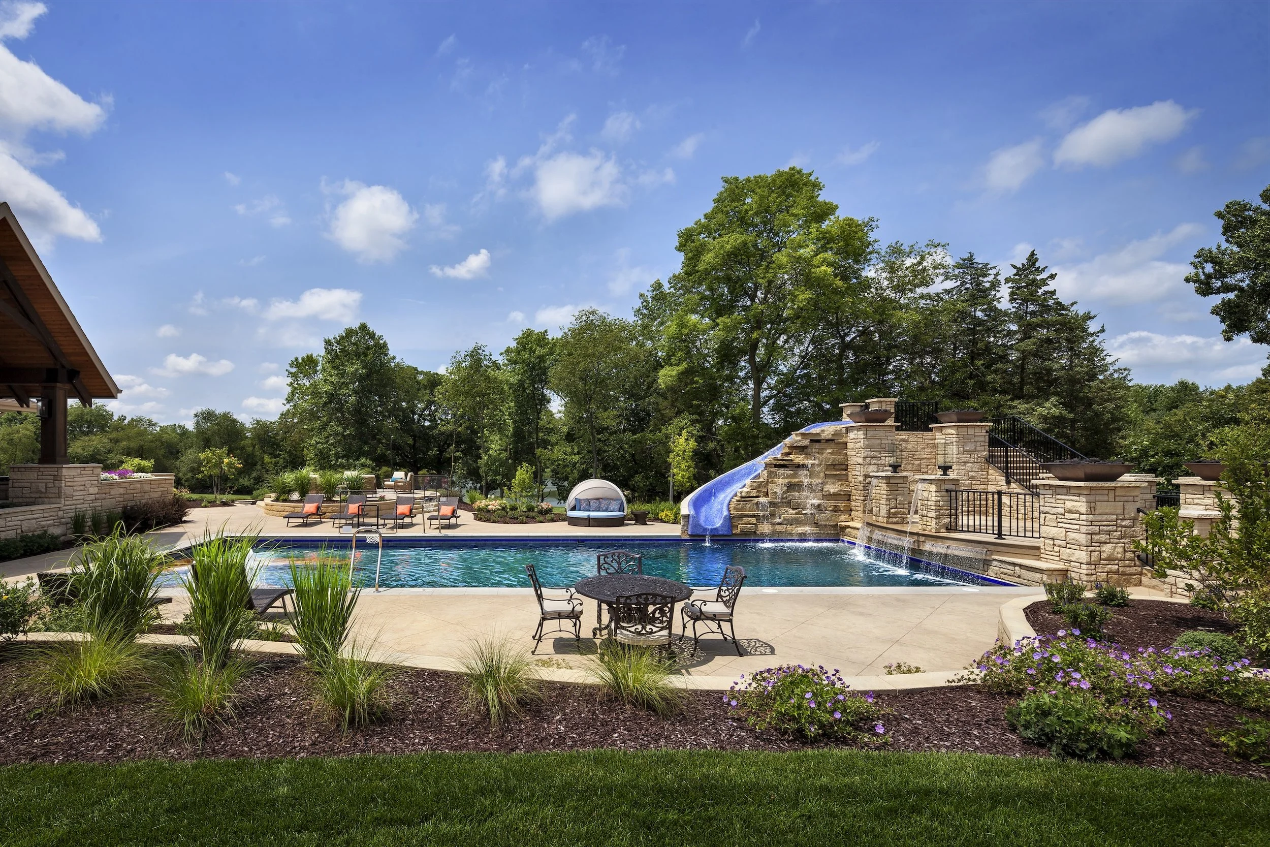 Luxurious outdoor swimming pool with waterfall, slide, and patio furniture surrounded by lush greenery and clear blue sky.