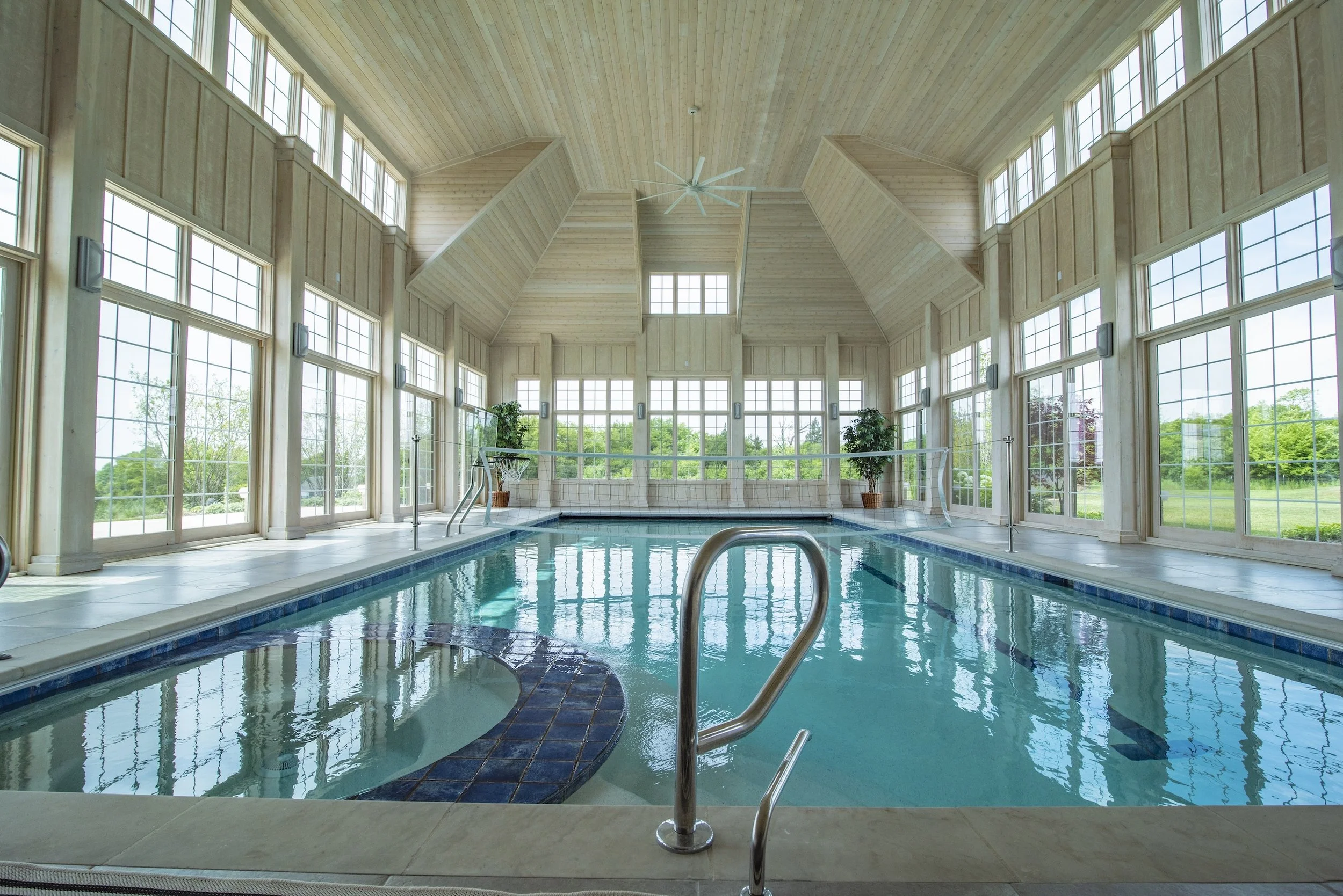 Luxurious indoor swimming pool with large windows and wood-paneled ceiling, featuring a built-in hot tub and hand railings.