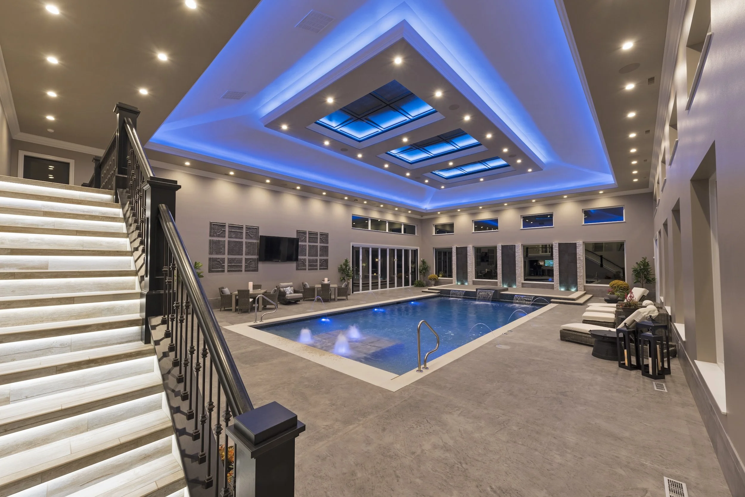 Luxury indoor swimming pool area with modern lighting, elegant staircase, and comfortable seating arrangements. The ceiling features recessed and blue accent lighting, enhancing the contemporary atmosphere.