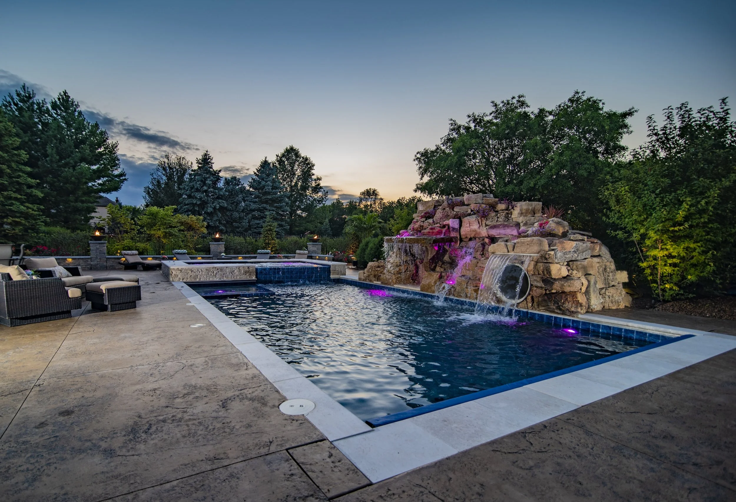 Outdoor swimming pool with waterfall feature, surrounded by patio furniture and greenery at dusk.