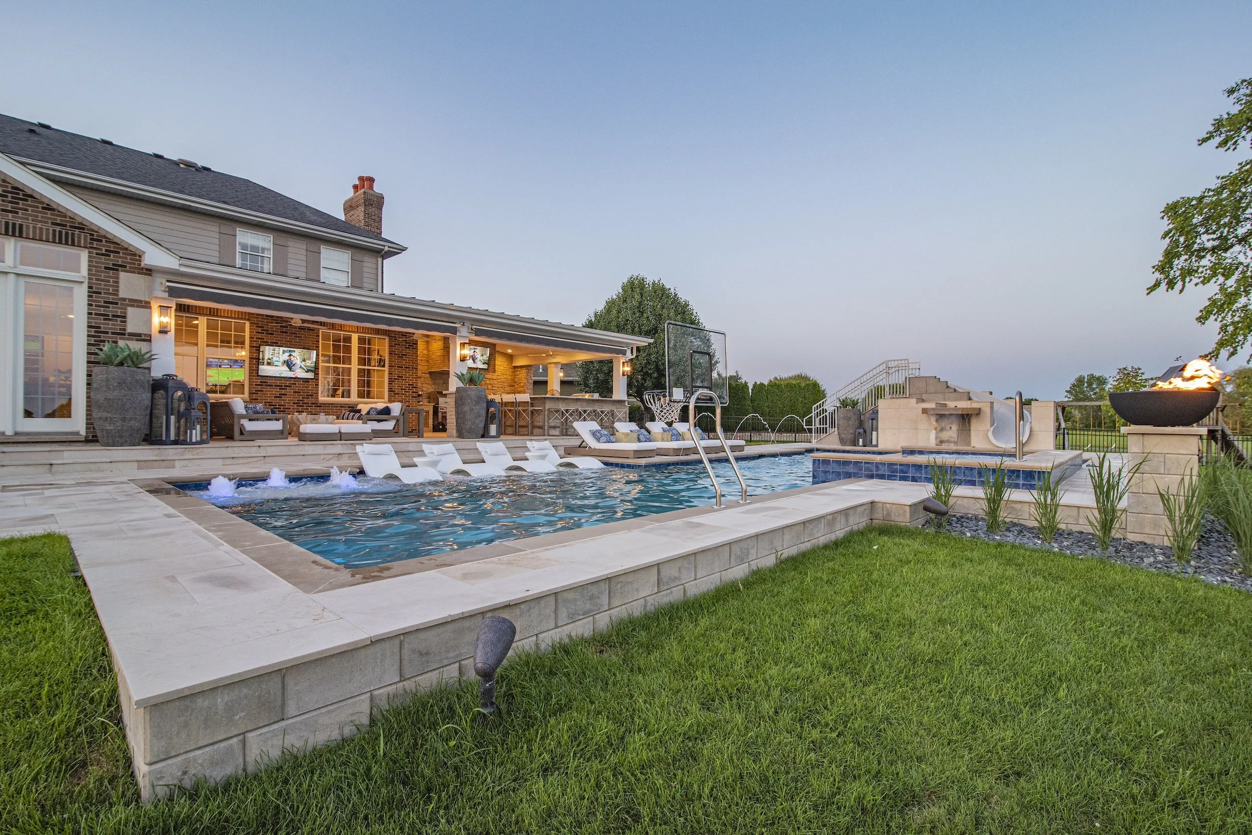 Luxurious backyard with a swimming pool featuring integrated lounge chairs, a poolside basketball hoop, a patio with outdoor seating, fire feature, and a brick house with a covered entertainment area.