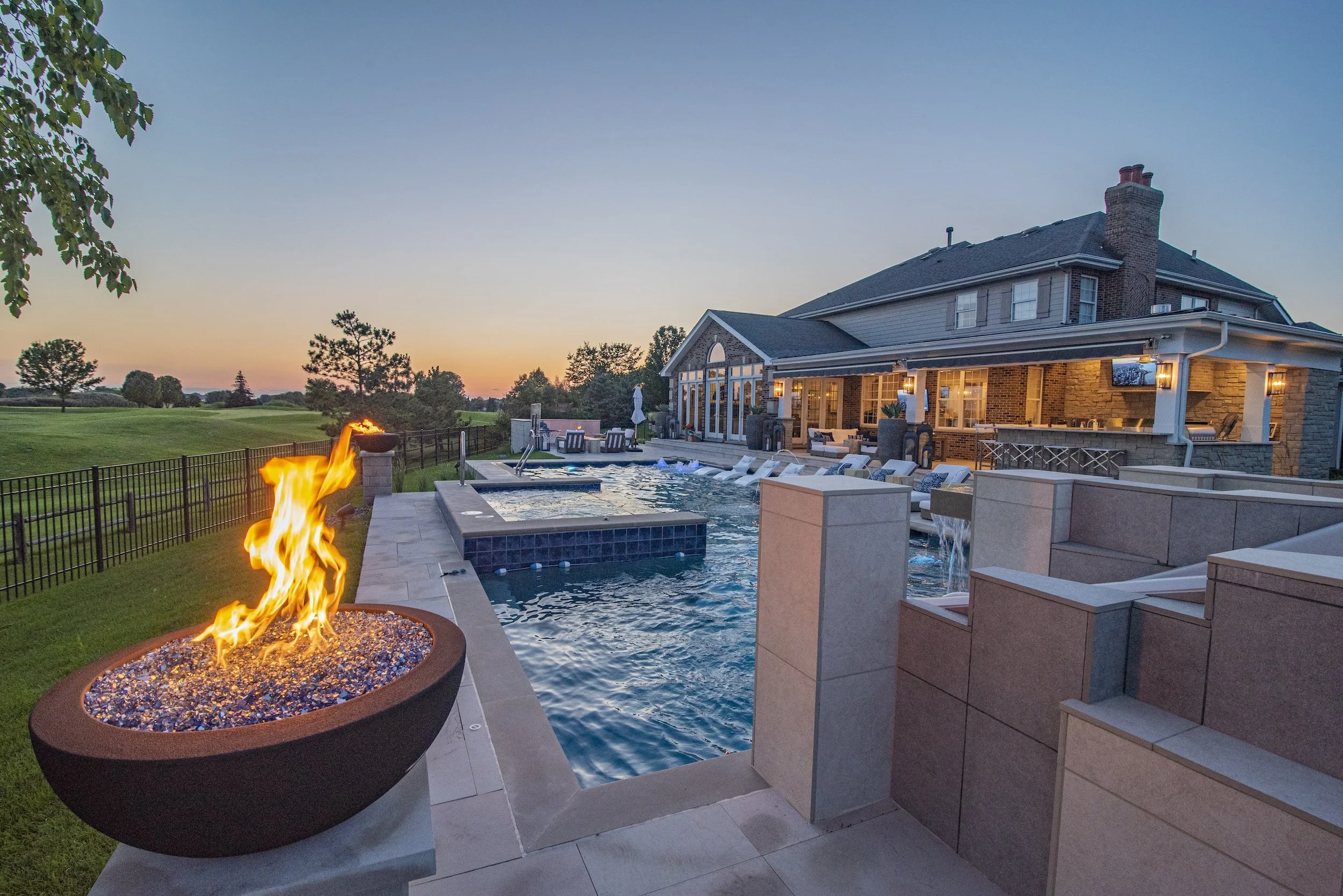 Luxury backyard with a swimming pool, fire pit, and lounge chairs beside a house at sunset.