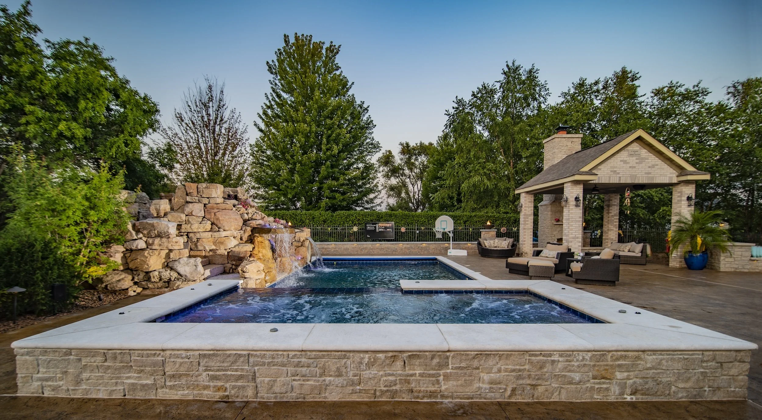 Backyard with a swimming pool and waterfall feature, surrounded by lush trees and landscaping. A pergola with seating and a fireplace is visible on the patio.