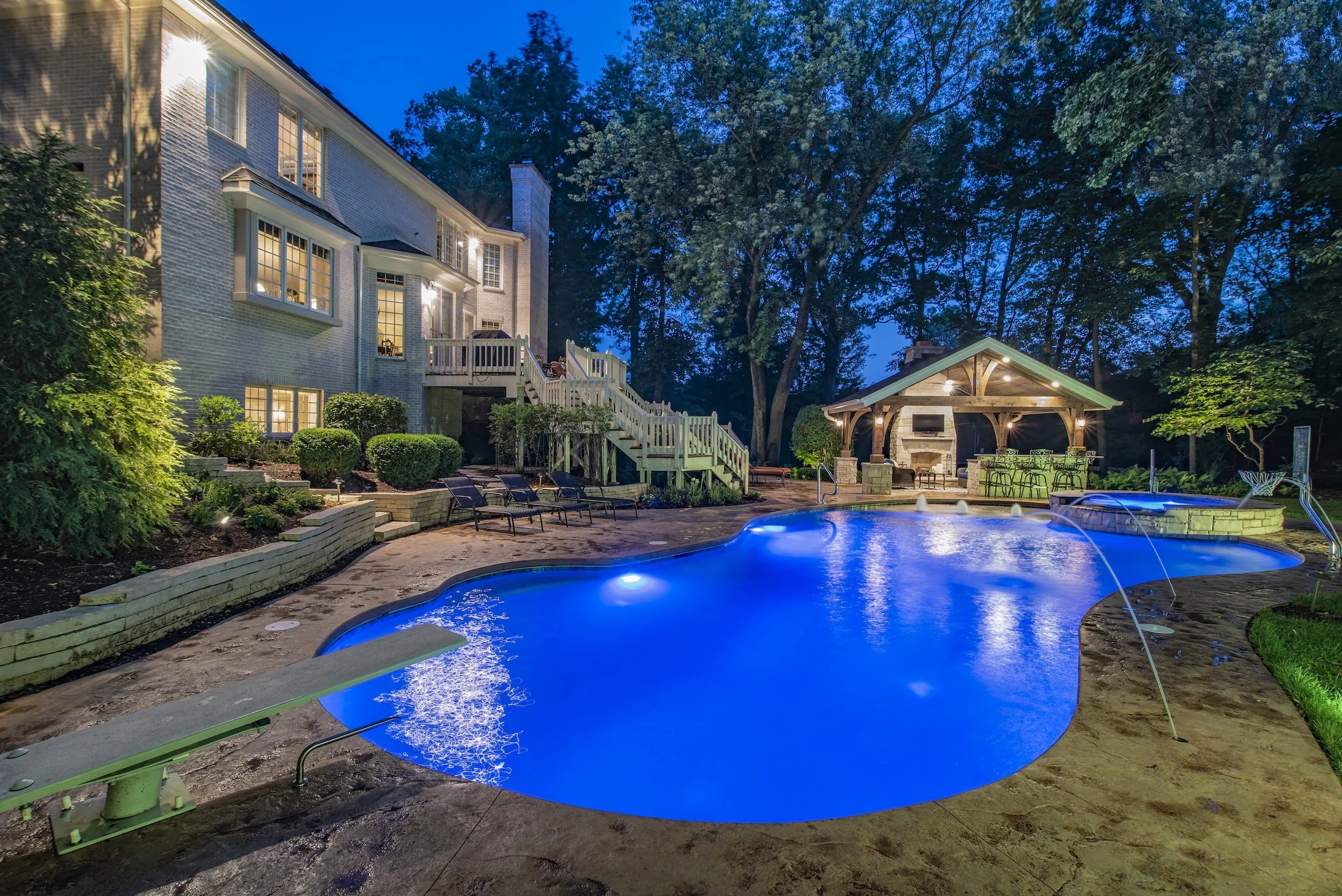 Luxurious backyard with illuminated swimming pool, diving board, patio, and gazebo in a wooded setting at dusk.