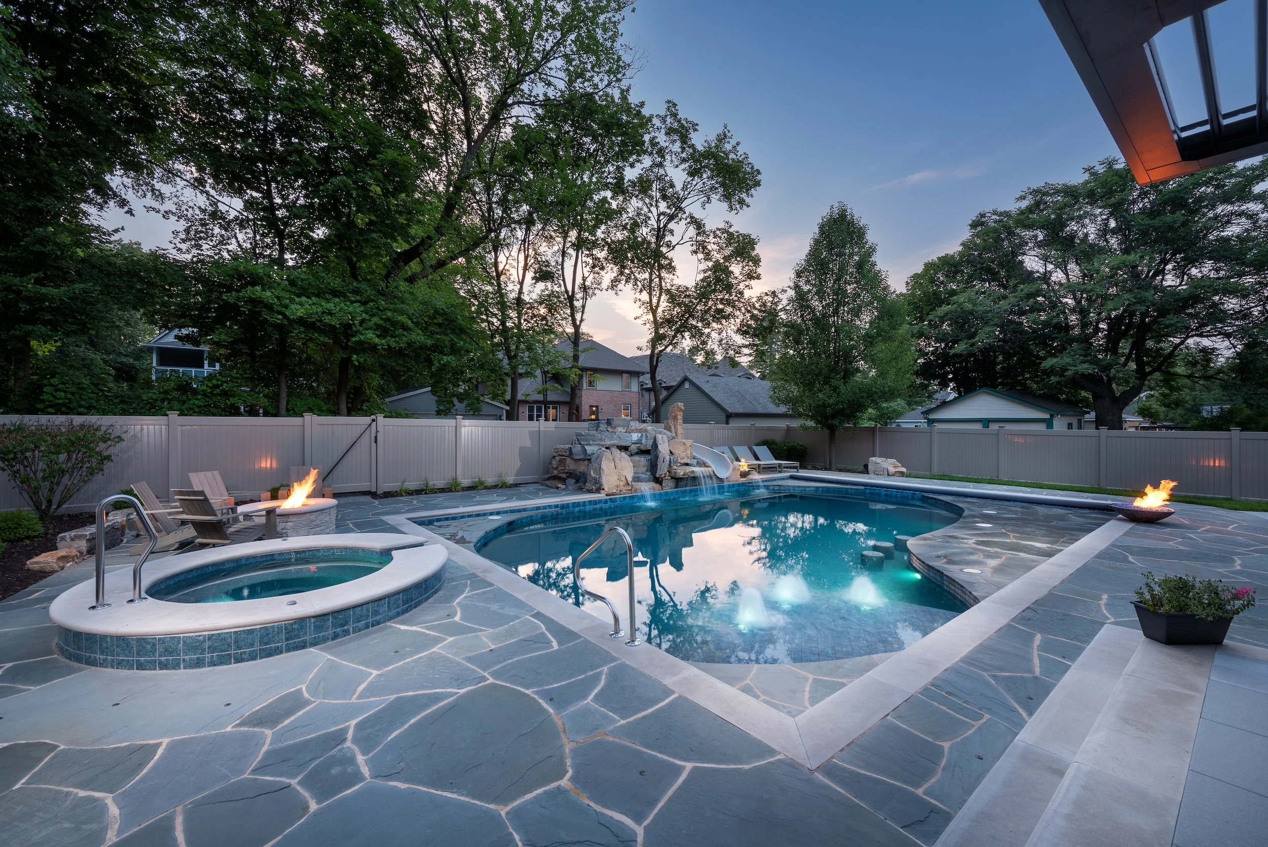 Backyard with a swimming pool, hot tub, fire pit, lounge chairs, and surrounding trees at dusk.