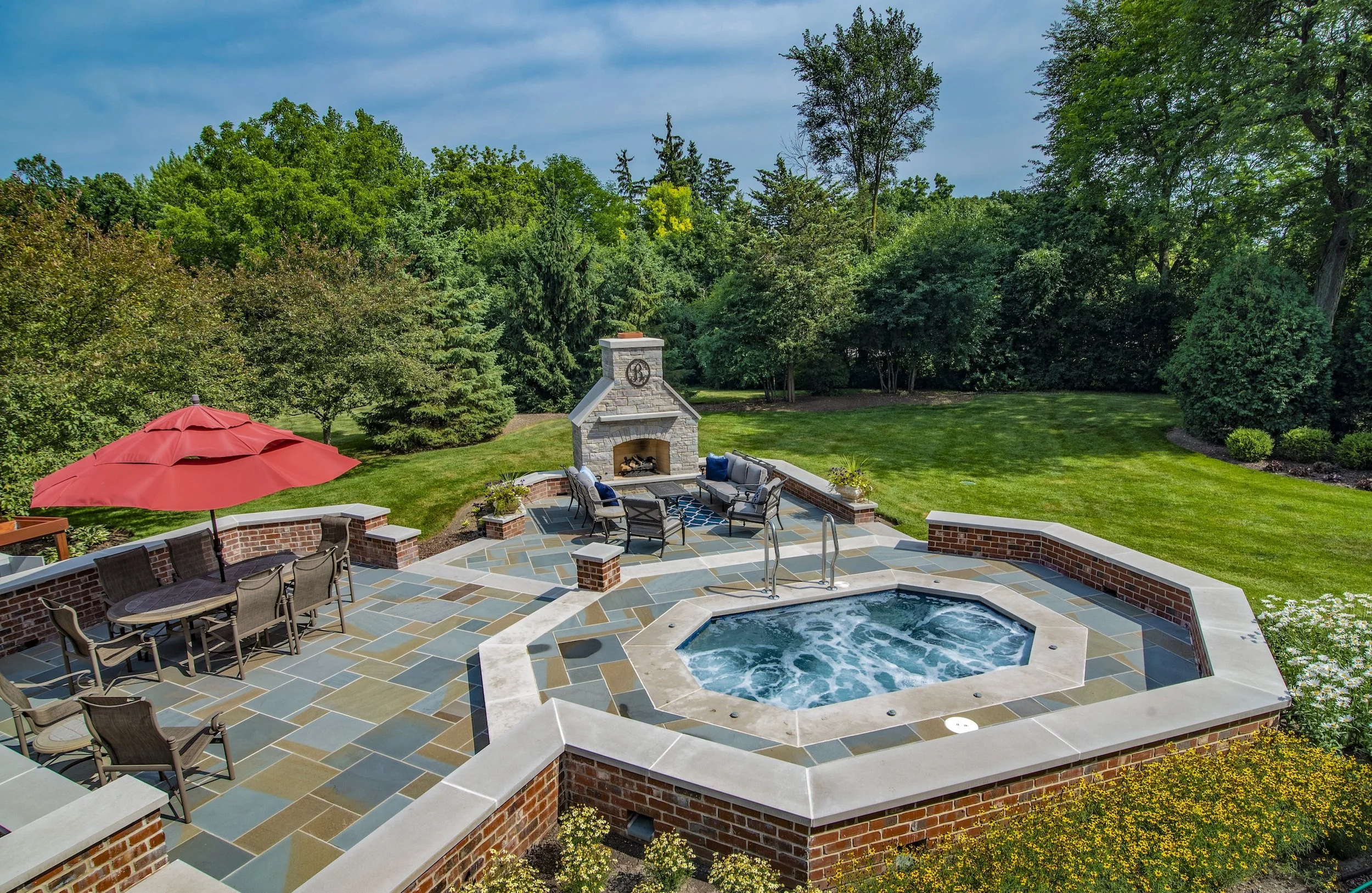 Backyard patio with a hot tub, outdoor seating, red umbrella, and fireplace surrounded by greenery.